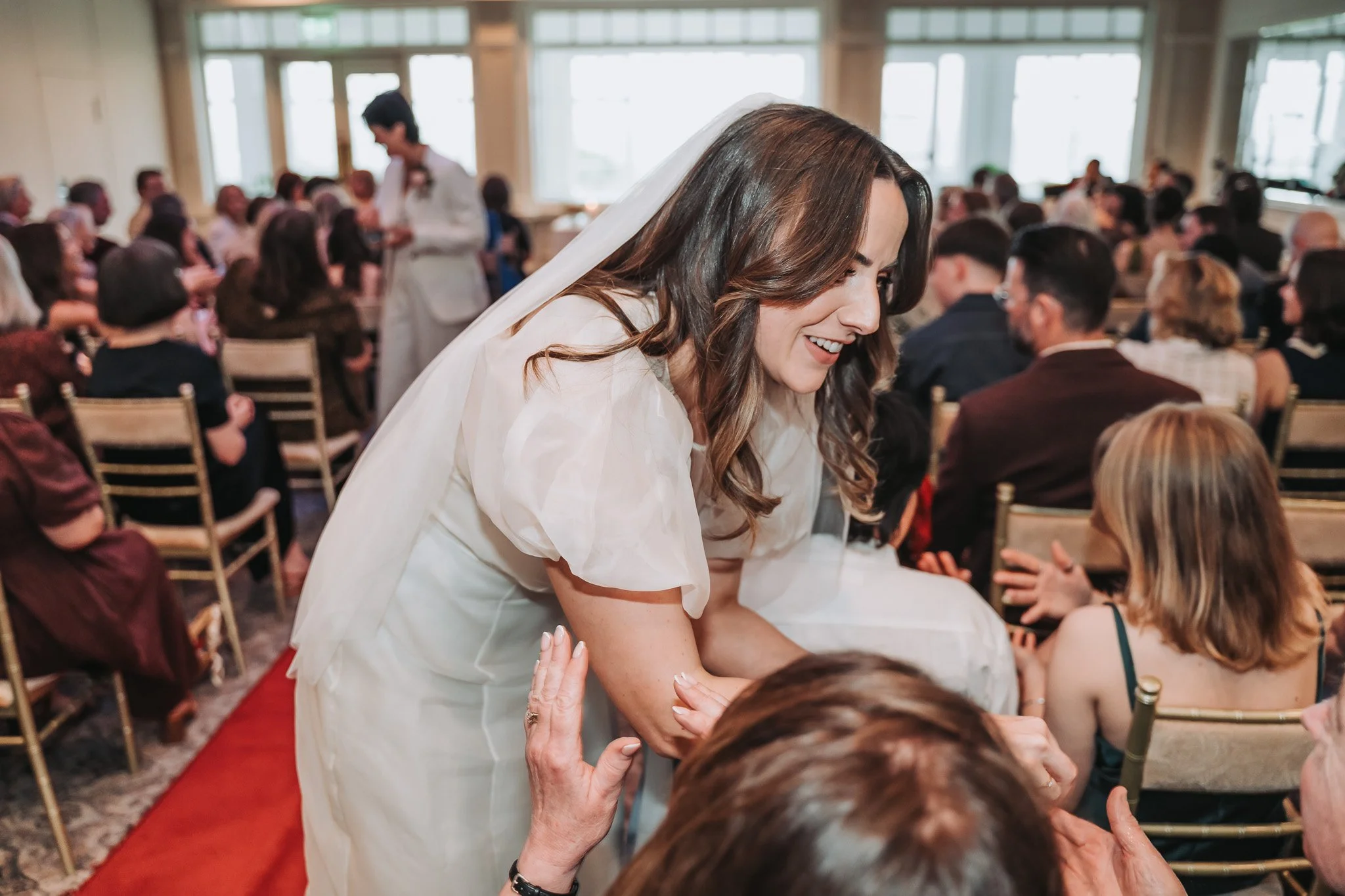 A woman with long brown hair dressed in a white dress and veil, smiling and shaking hands with guests at her wedding reception in a well-lit room filled with seated guests.