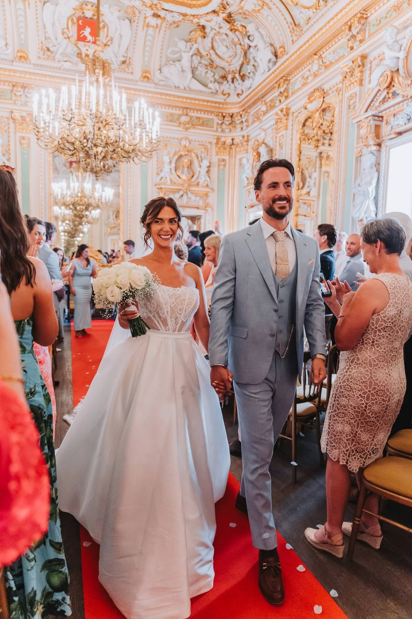 A newlywed couple walking hand in hand down a red carpet aisle in an ornately decorated hall, surrounded by wedding guests. The bride is wearing a wedding gown and holding a bouquet of white roses. The groom is dressed in a light gray suit. The hall 