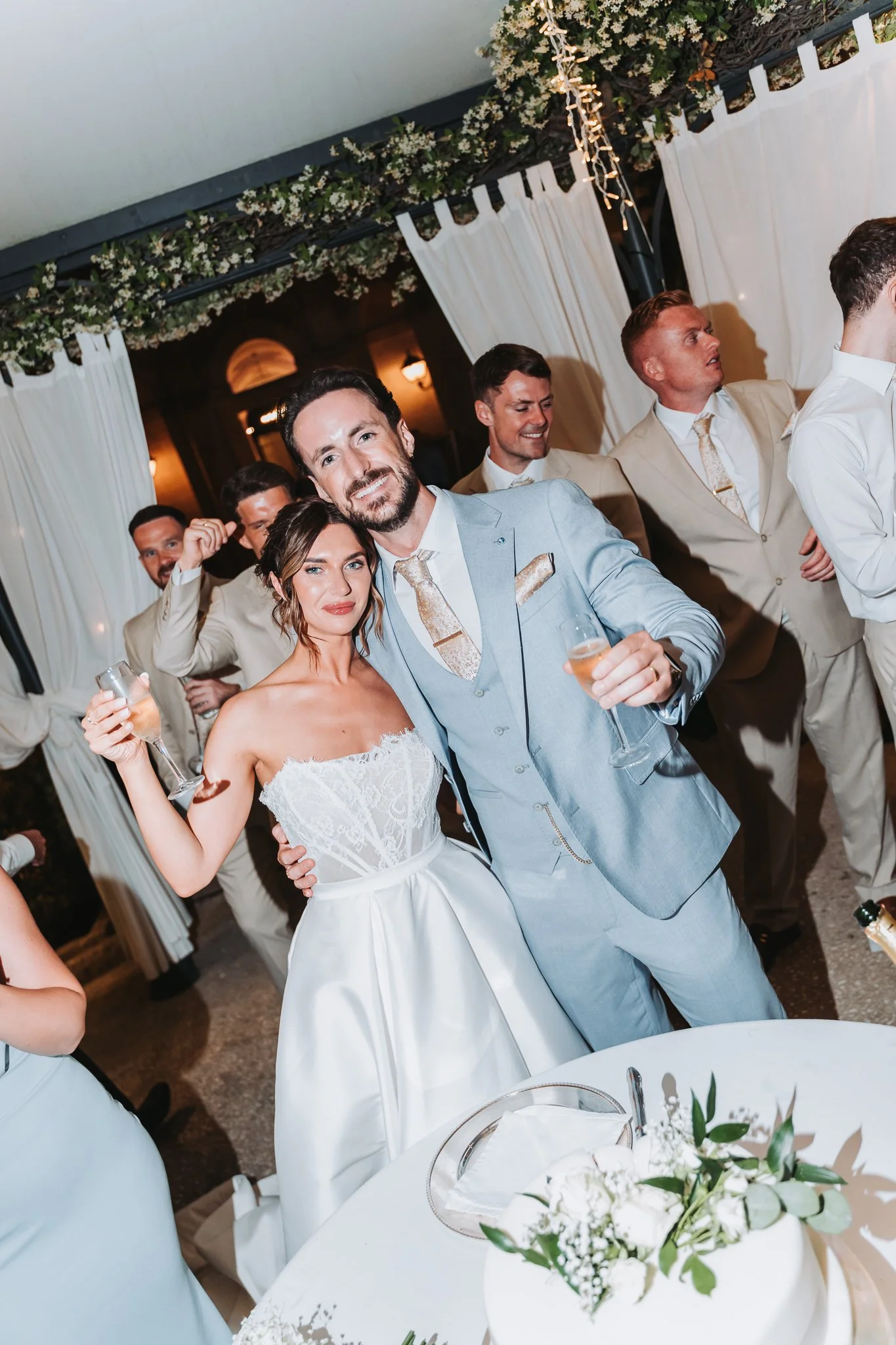 Bride and groom at their wedding reception with friends, holding champagne glasses, surrounded by floral decor and white curtains.