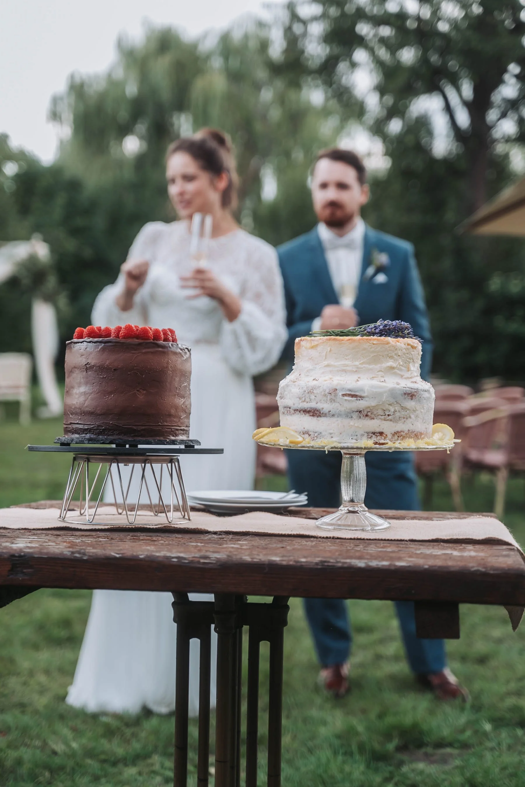 Two cakes, one chocolate with raspberries and one semi-naked with lavender, on a rustic wooden table outdoors, with blurred people in the background celebrating.