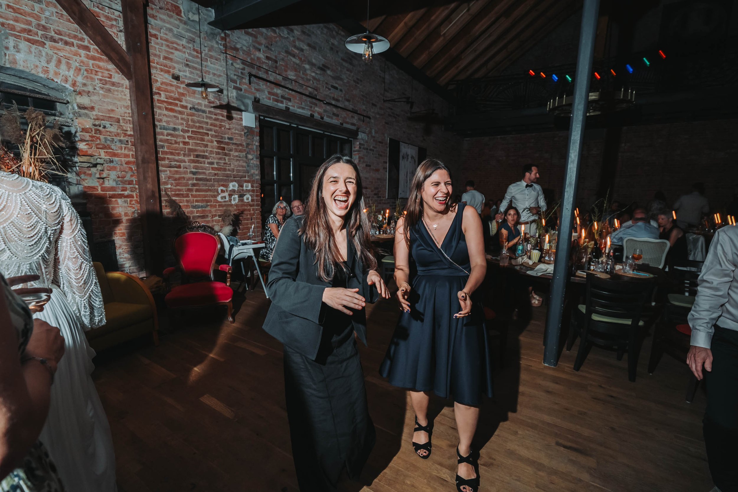 Two women laughing and smiling while dancing at a party in a venue with exposed brick walls, wooden beams, and dim lighting. They are dressed in elegant black outfits.
