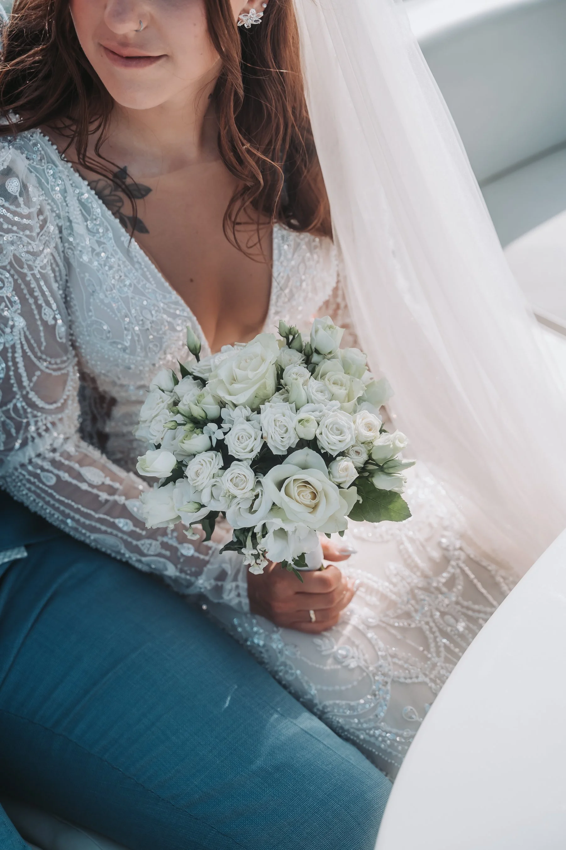 A woman in a wedding dress holding a bouquet of white roses, sitting next to another woman in a blue outfit. She is wearing earrings, a nose ring, and has a tattoo on her chest. There is a white veil in the background.
