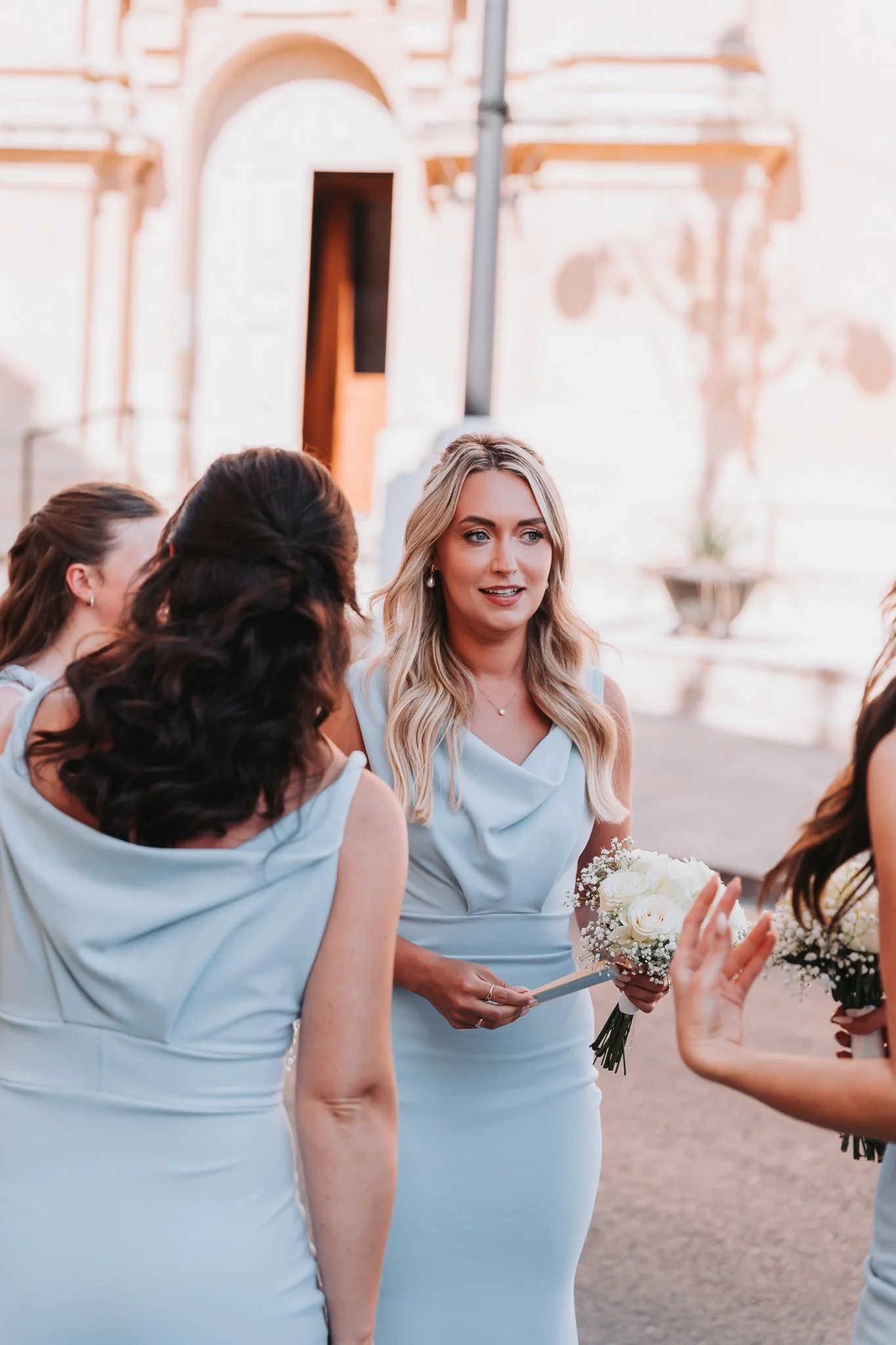 A group of women in light blue dresses gathered outdoors, with one woman holding a bouquet of white flowers, during what appears to be a wedding or special event.
