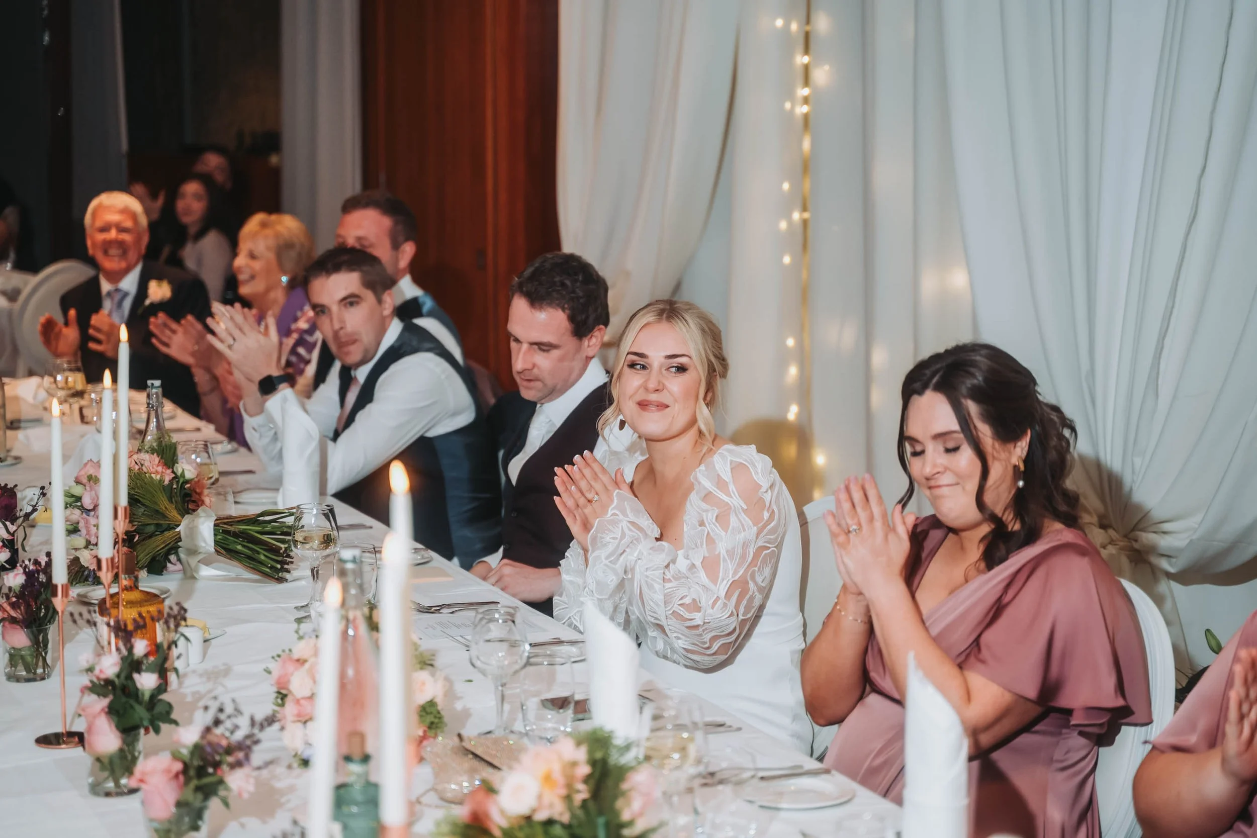 People sitting at a decorated table during a wedding reception, with floral centerpieces and candles.