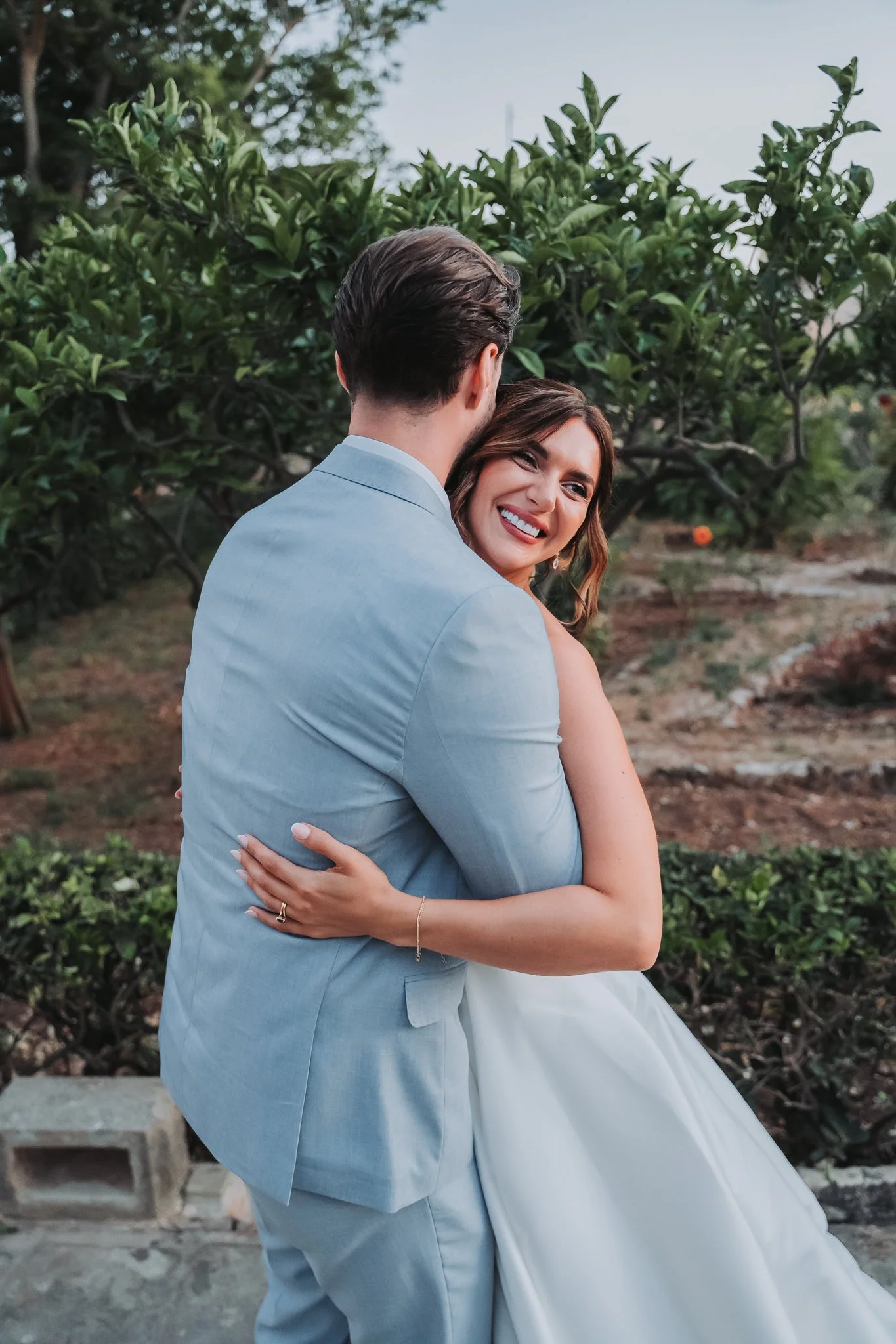 A couple hugging outdoors, man in a light blue suit, woman in a white dress, smiling with eyes closed near green foliage.