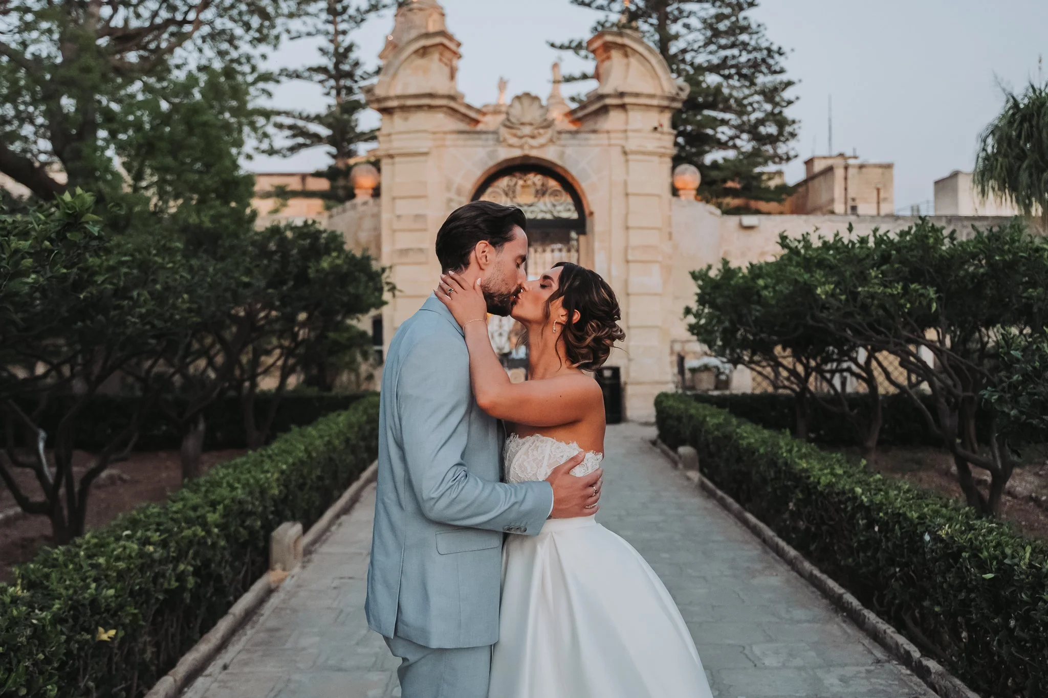 A couple in wedding attire sharing a kiss outdoors with a historic arched gate and trees behind them.
