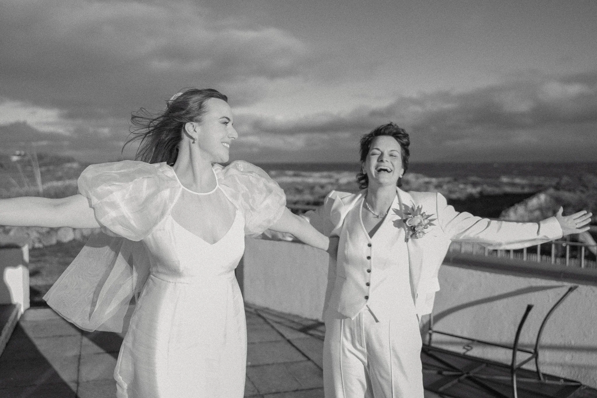 Two women with outstretched arms smiling and enjoying outdoor view, with a cloudy sky and railing in the background.