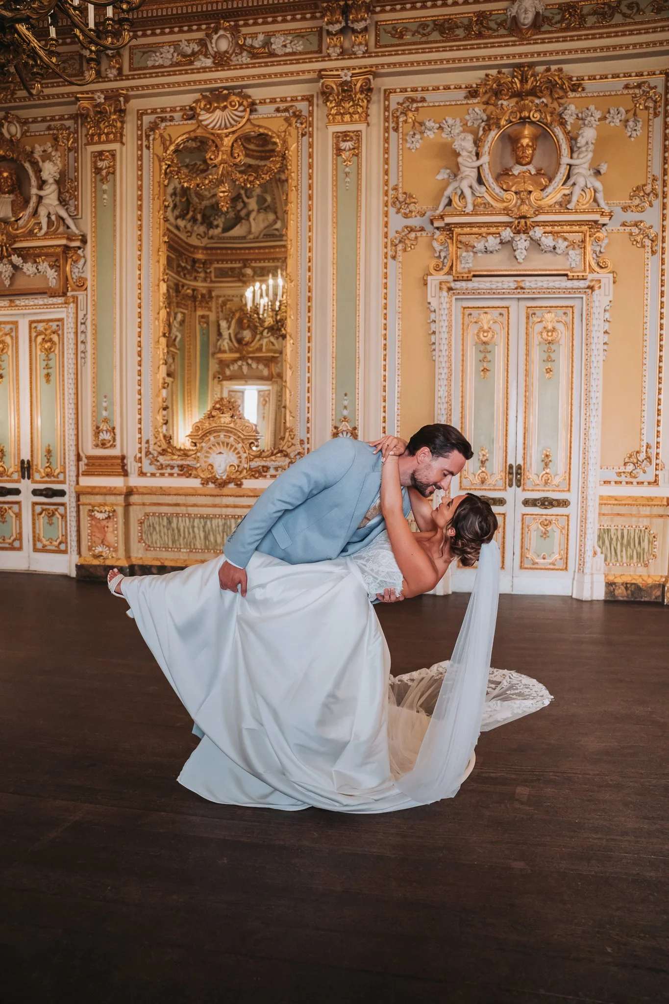 A bride and groom are dancing in a luxurious, ornately decorated room with gold accents, intricate moldings, and a large mirror. The groom is lifting the bride, who is wearing a white wedding dress with lace details, and they are gazing into each oth