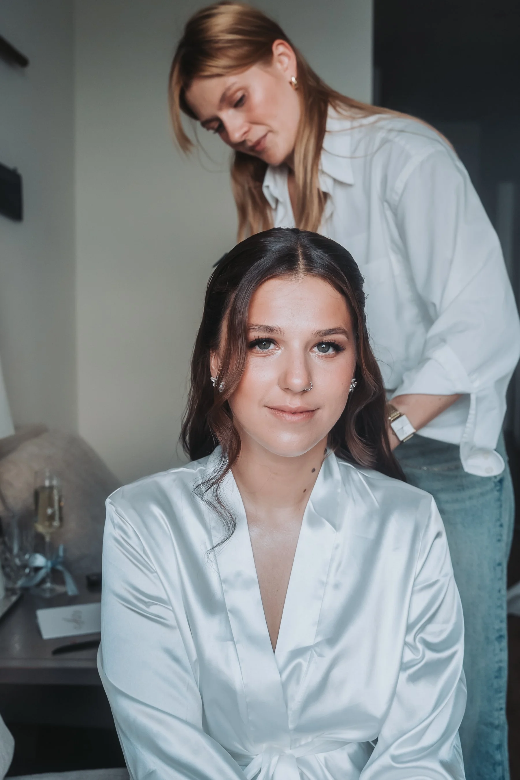 A young woman with brown hair and fair skin sitting in a studio while a hairdresser styles her hair. She is wearing a white satin robe and has a calm expression.