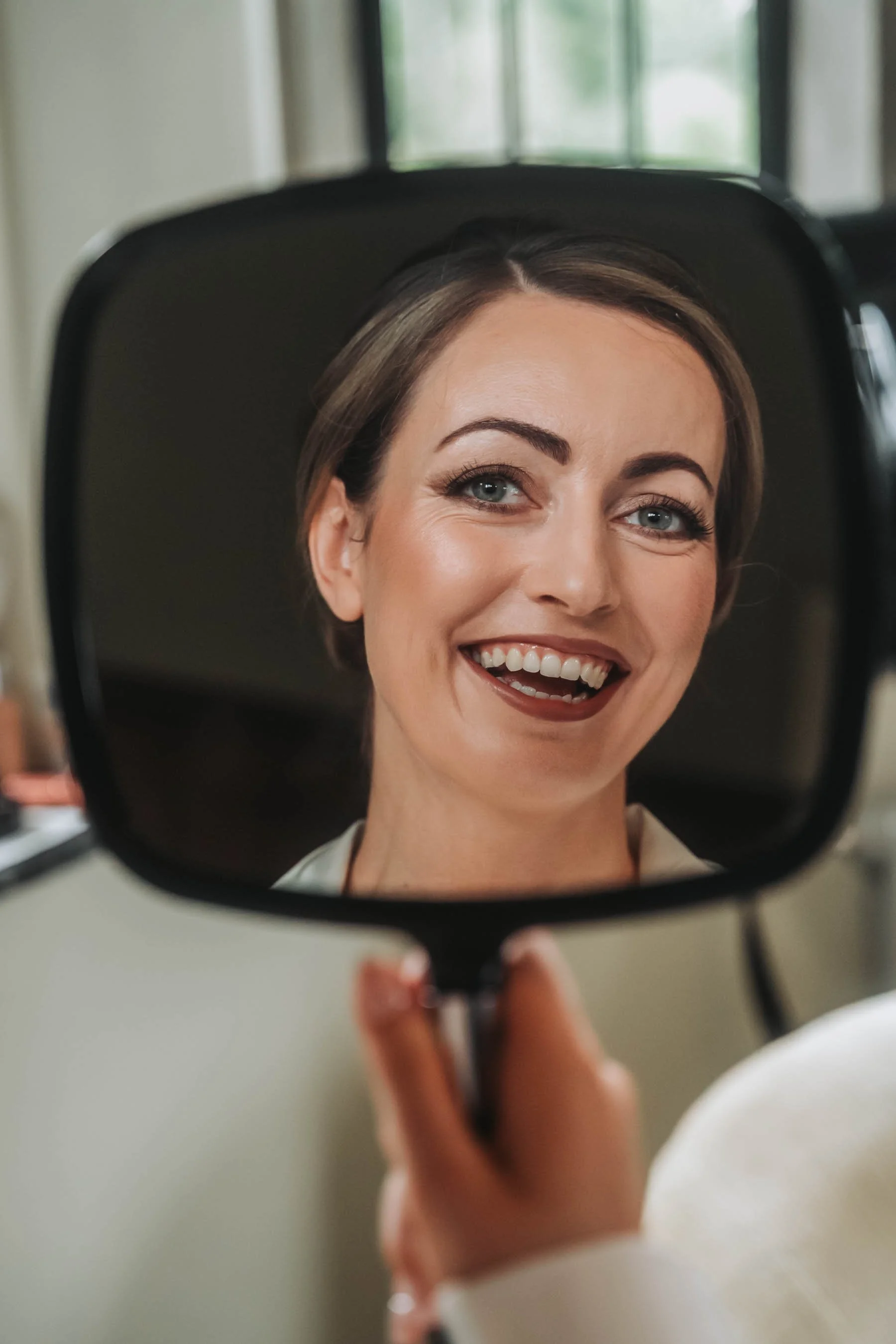 A woman smiling and looking into a mirror, with her reflection visible. The photo is taken through the mirror, showing her face and upper shoulders.
