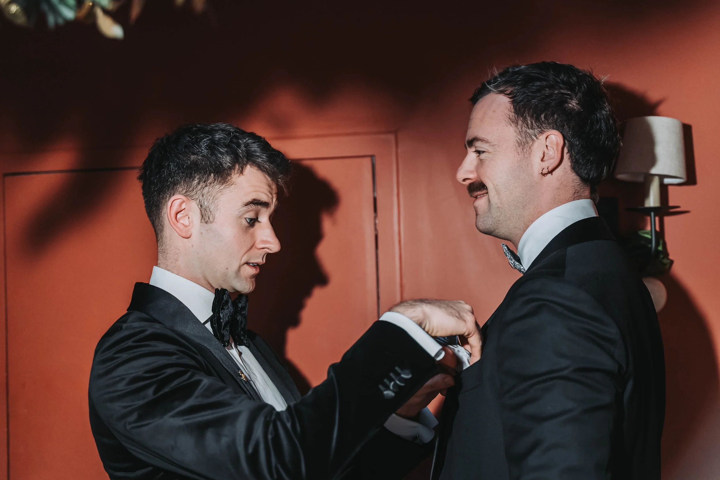 Two men dressed in tuxedos facing each other, holding hands and smiling, in a warm-hued room, possibly at a formal event or wedding.