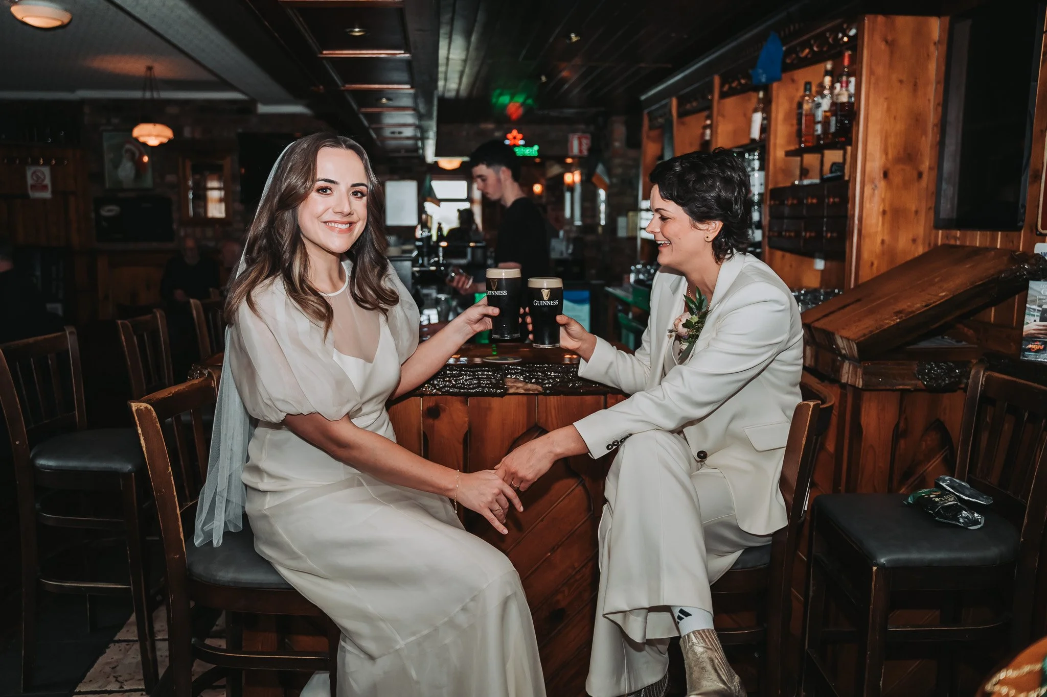 Two women dressed in wedding attire sitting in a bar, smiling and clinking Guinness glasses. One woman has long, wavy brown hair and the other has short, curly black hair. They are holding hands and appear to be celebrating.
