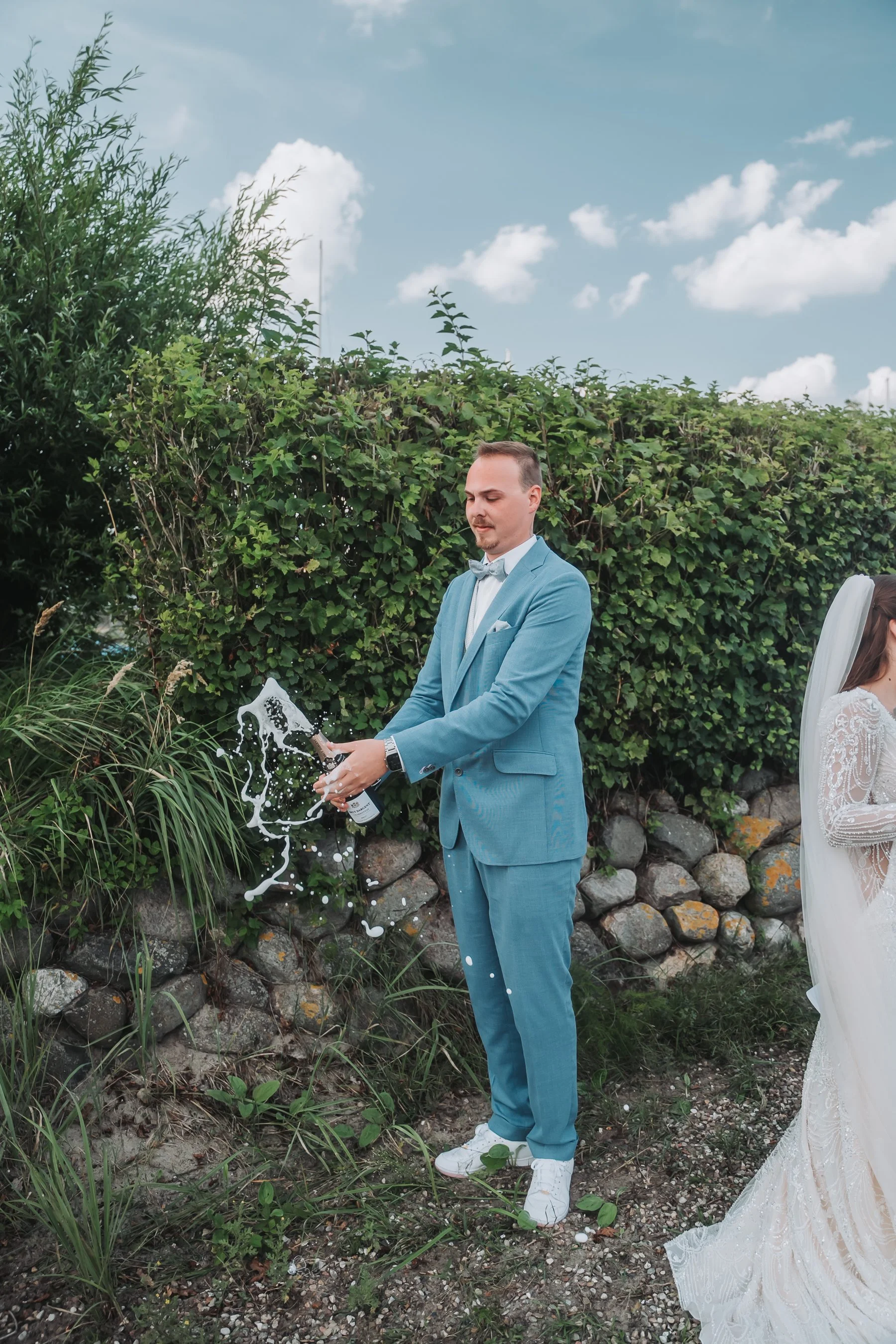 A man in a blue suit with a white shirt and gray bow tie pouring champagne outdoors, with greenery and rocks behind him, and a woman in a white lace wedding dress partially visible on the right.