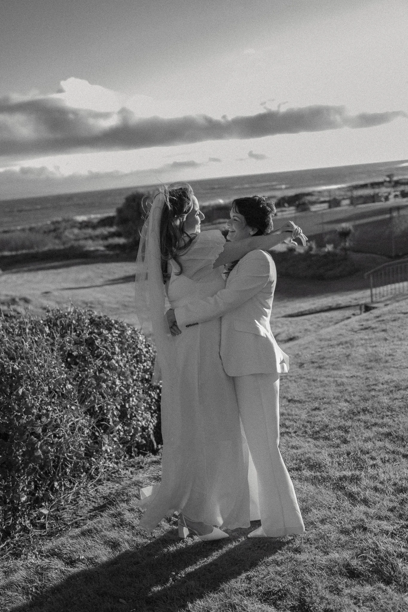 Black and white photo of a bride and groom embracing outdoors, with the ocean and cloudy sky in the background.