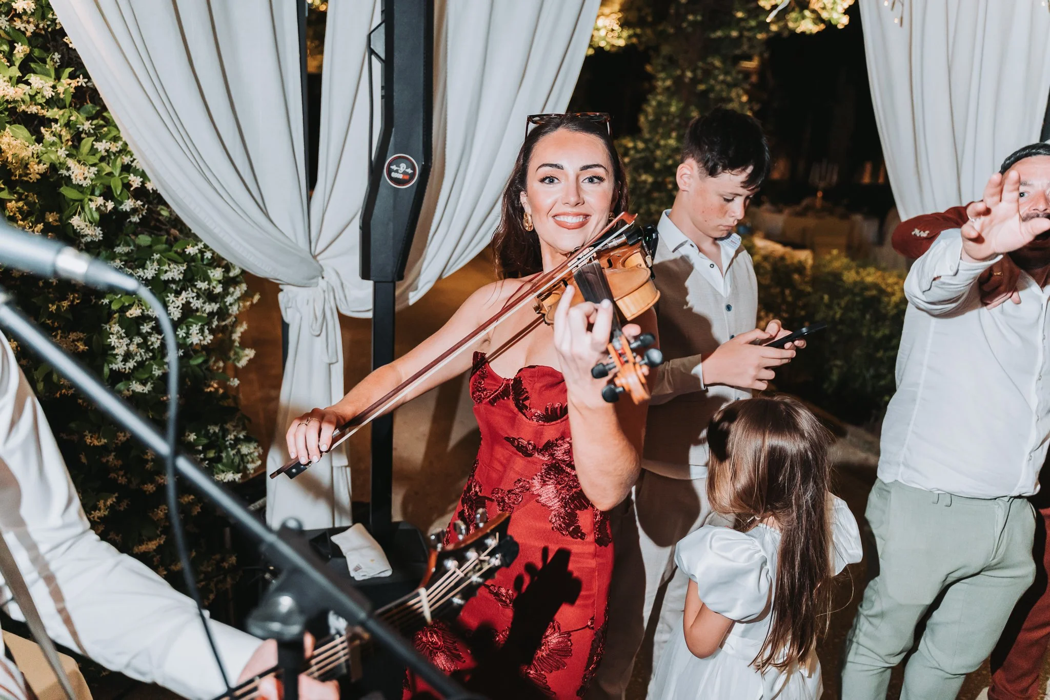 A woman in a red dress playing the violin at an outdoor gathering with other people around at night.