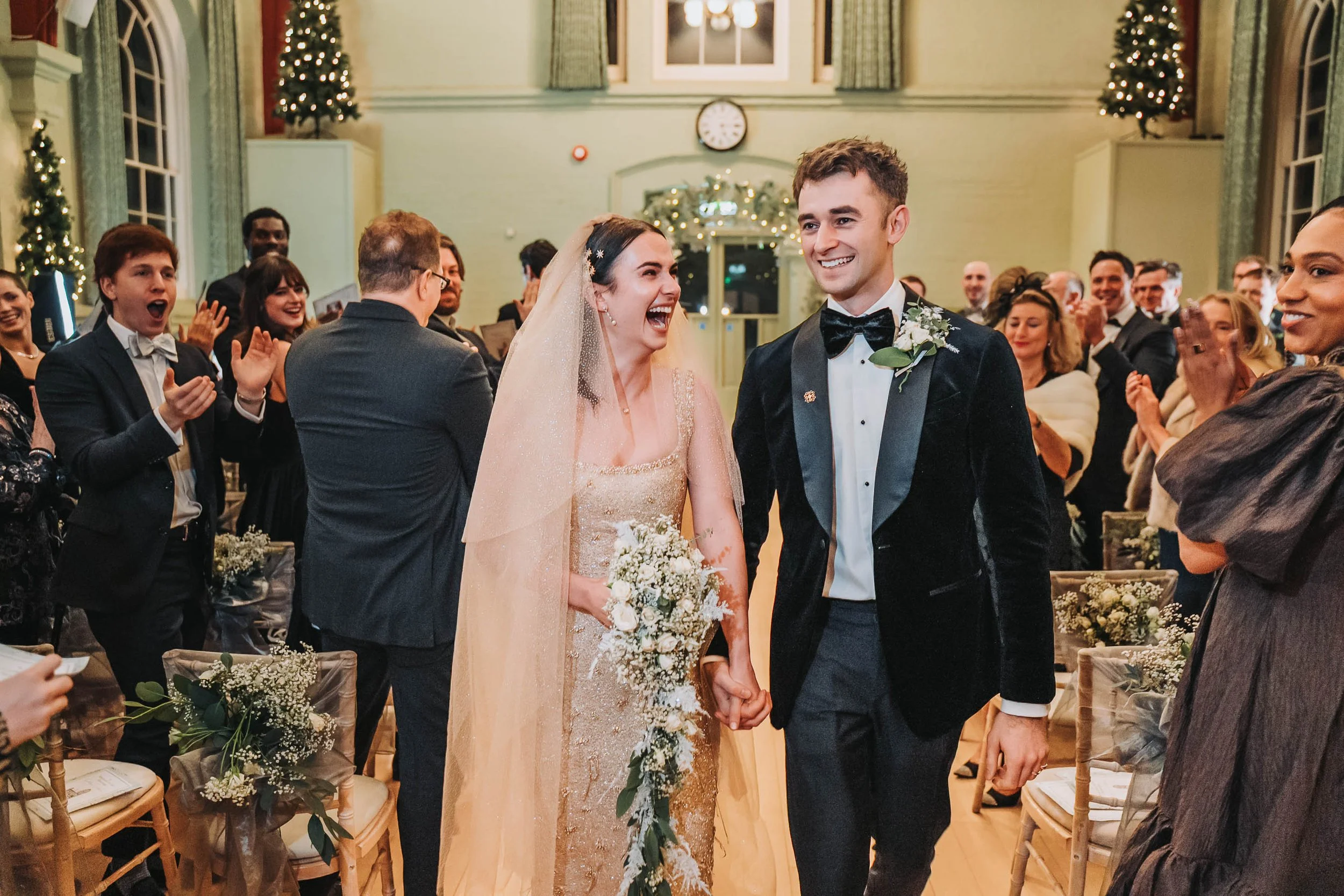 Young couple, bride in a gold dress with a veil, groom in a tuxedo, holding hands and smiling at each other during a wedding ceremony in a decorated hall with guests clapping and celebrating.