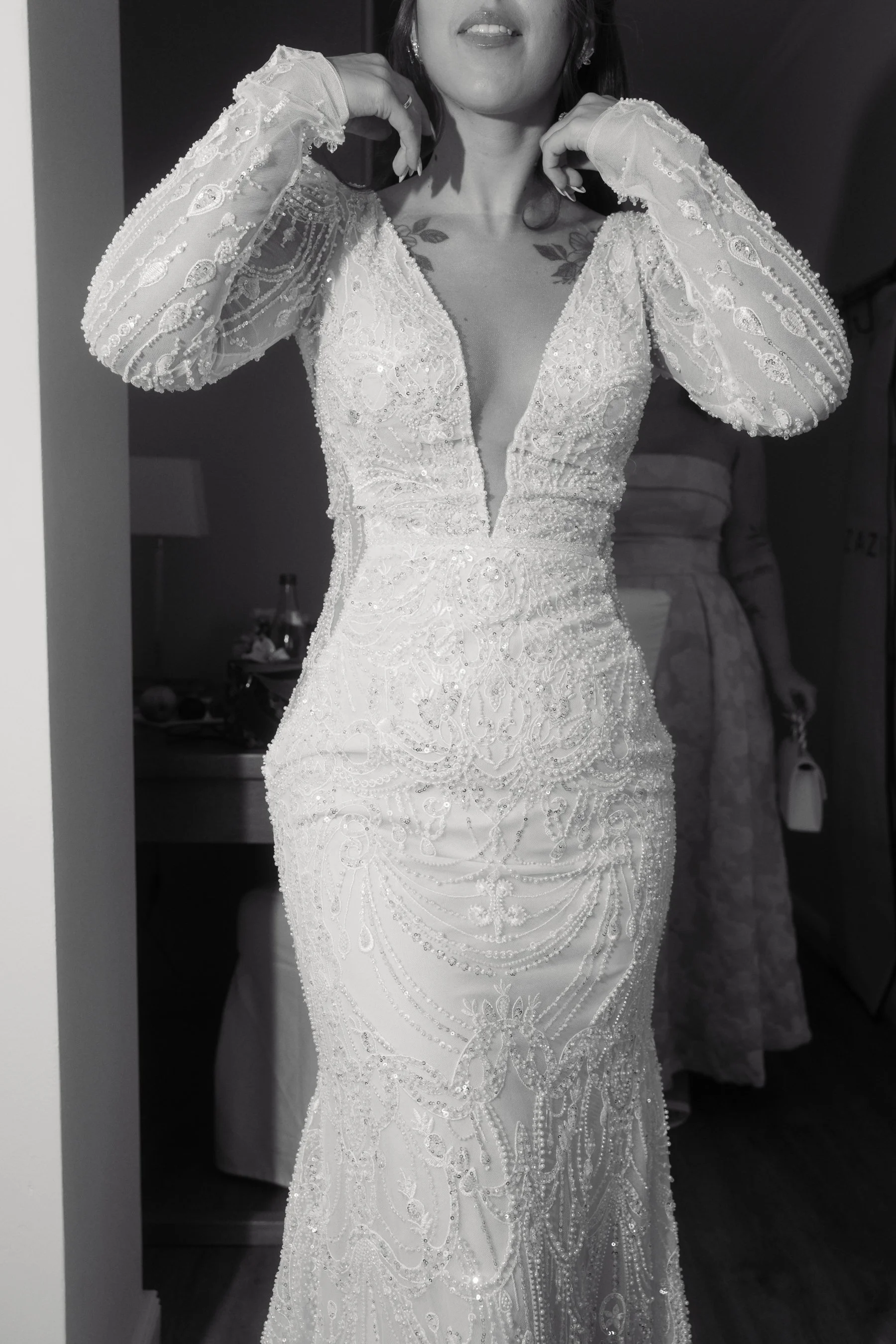 A woman trying on an ornate, fitted wedding gown with long sheer, embroidered sleeves, intricate bead and sequin embellishments, and a plunging neckline. She is adjusting the collar of the dress near her face.