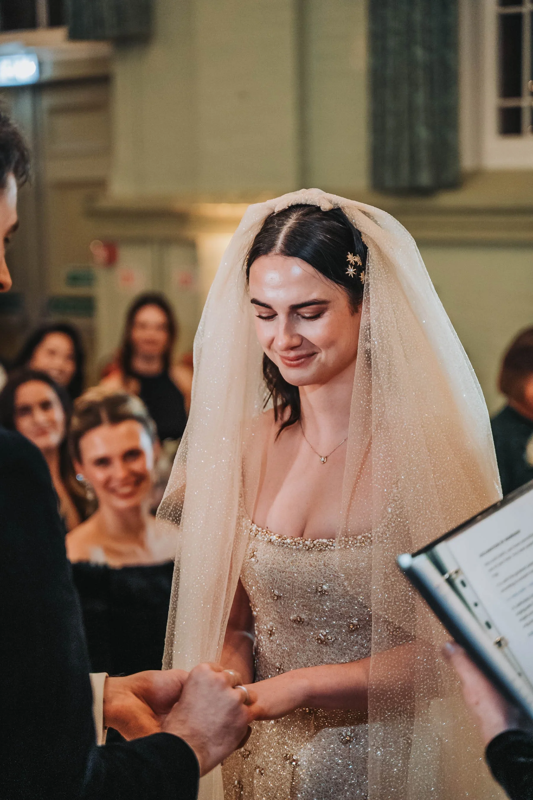 Bride with dark hair wearing a sparkly gold dress and veil, smiling during wedding ceremony, holding hands with groom, surrounded by guests.