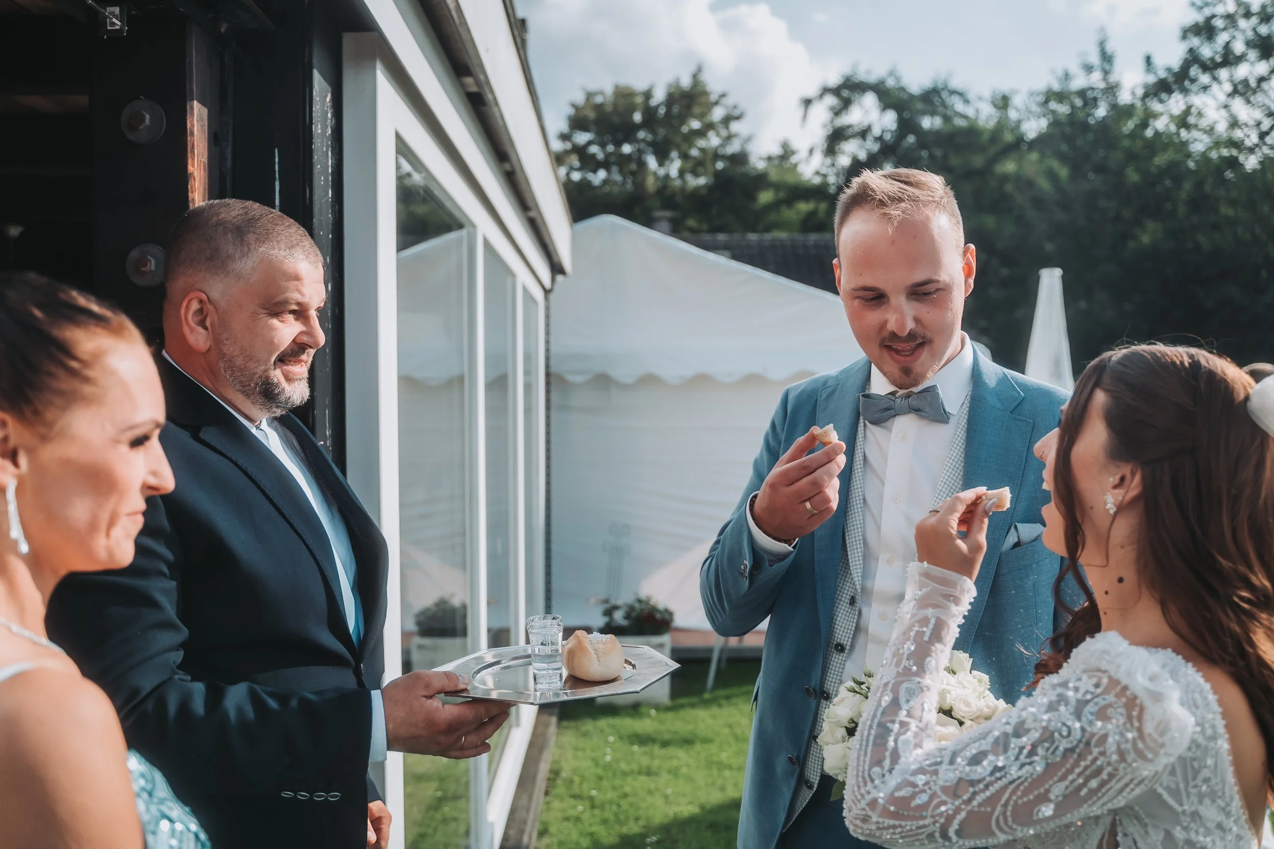 A wedding reception scene outdoors shows a man in a blue suit and bow tie holding a piece of bread and talking with a woman in a white lace dress with dark hair. A server in a black suit is holding a tray with bread rolls. Other guests are visible, a