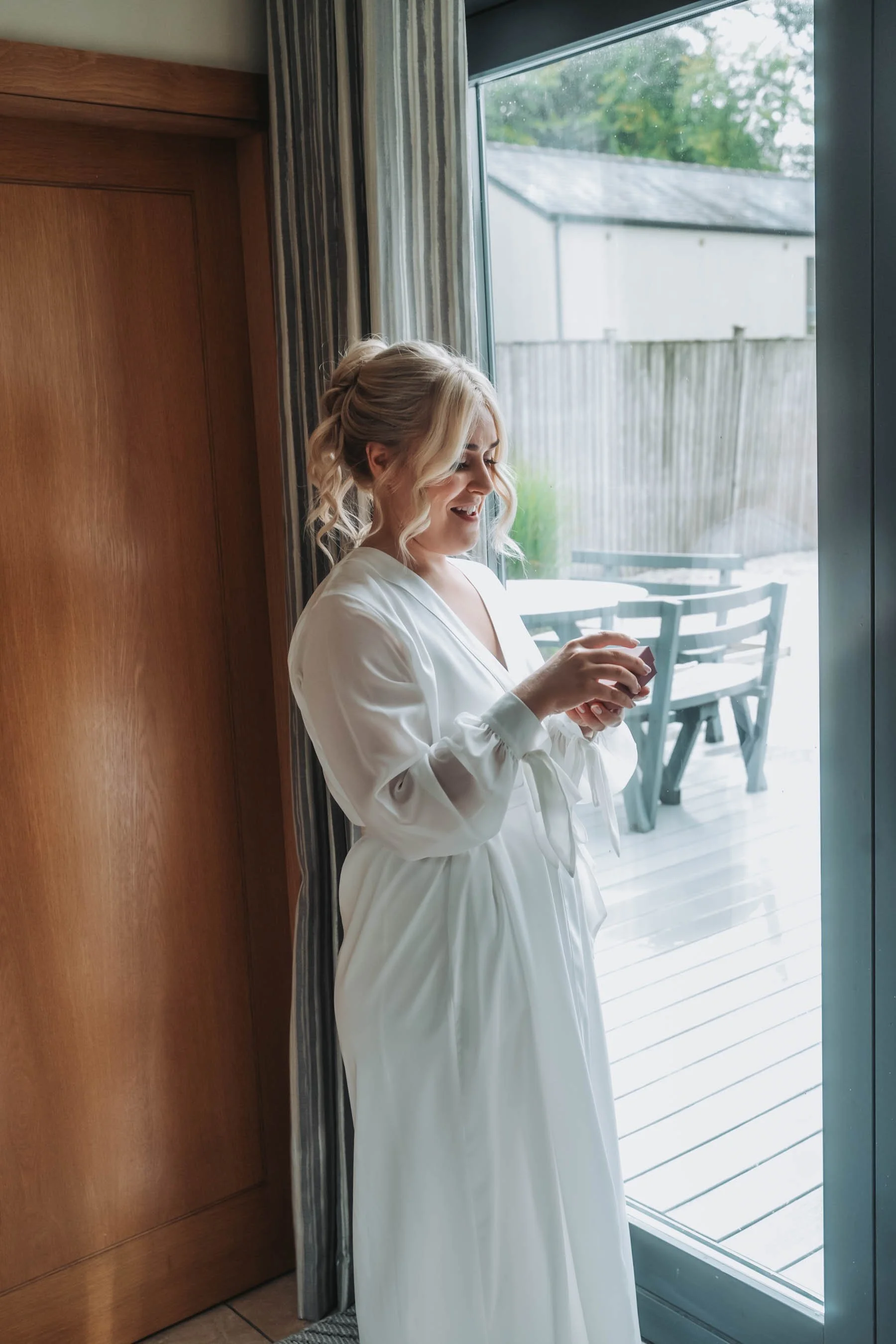 A woman with blonde, curly hair in a loose bun, standing by a large window, smiling and holding a small object in her hands. She is wearing a white robe and is next to a wooden door with a curtain partially drawn across the window. Outside, there is 