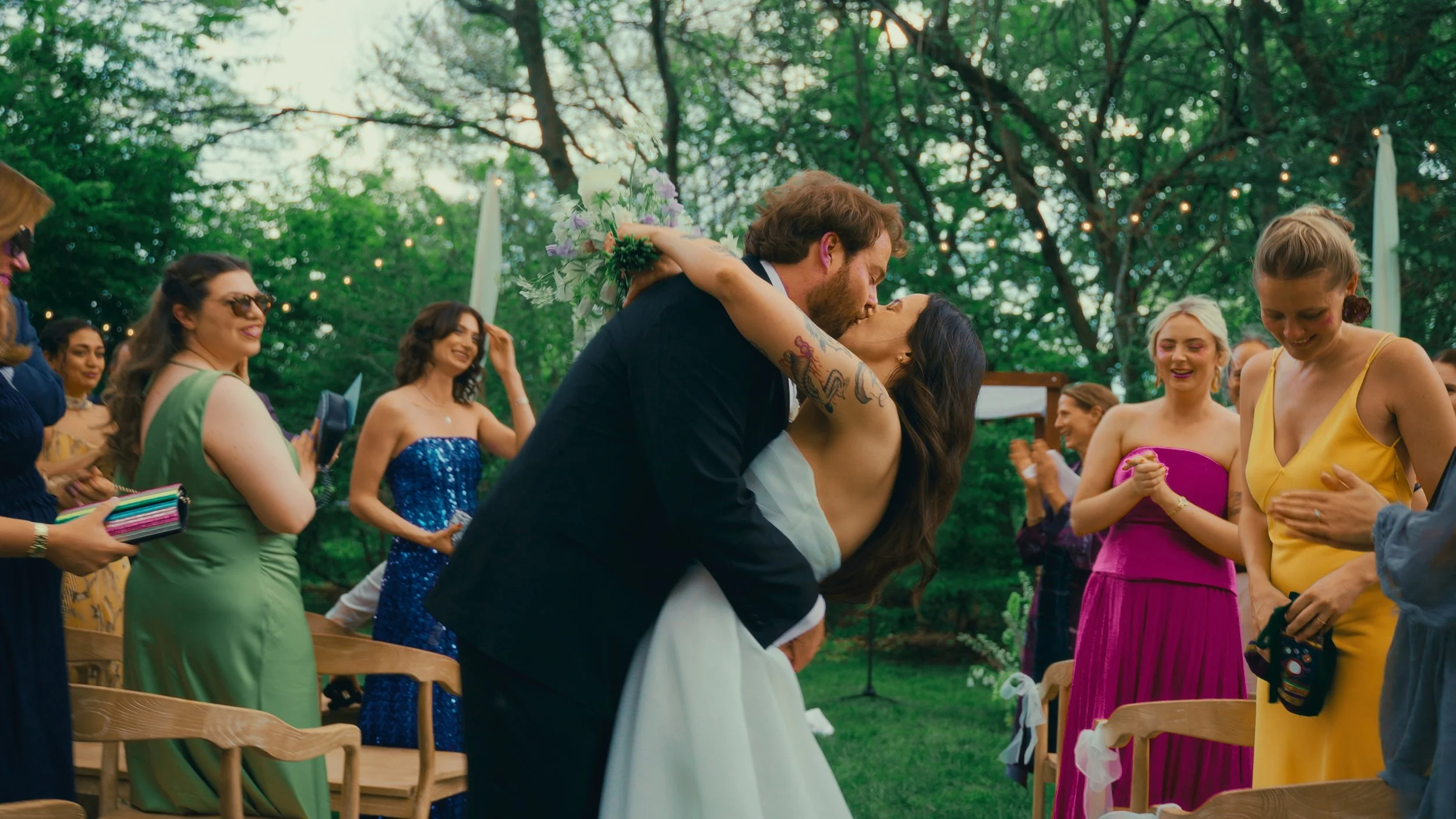 A couple kissing at an outdoor wedding ceremony, surrounded by wedding guests in colorful dresses, with green trees and string lights in the background.