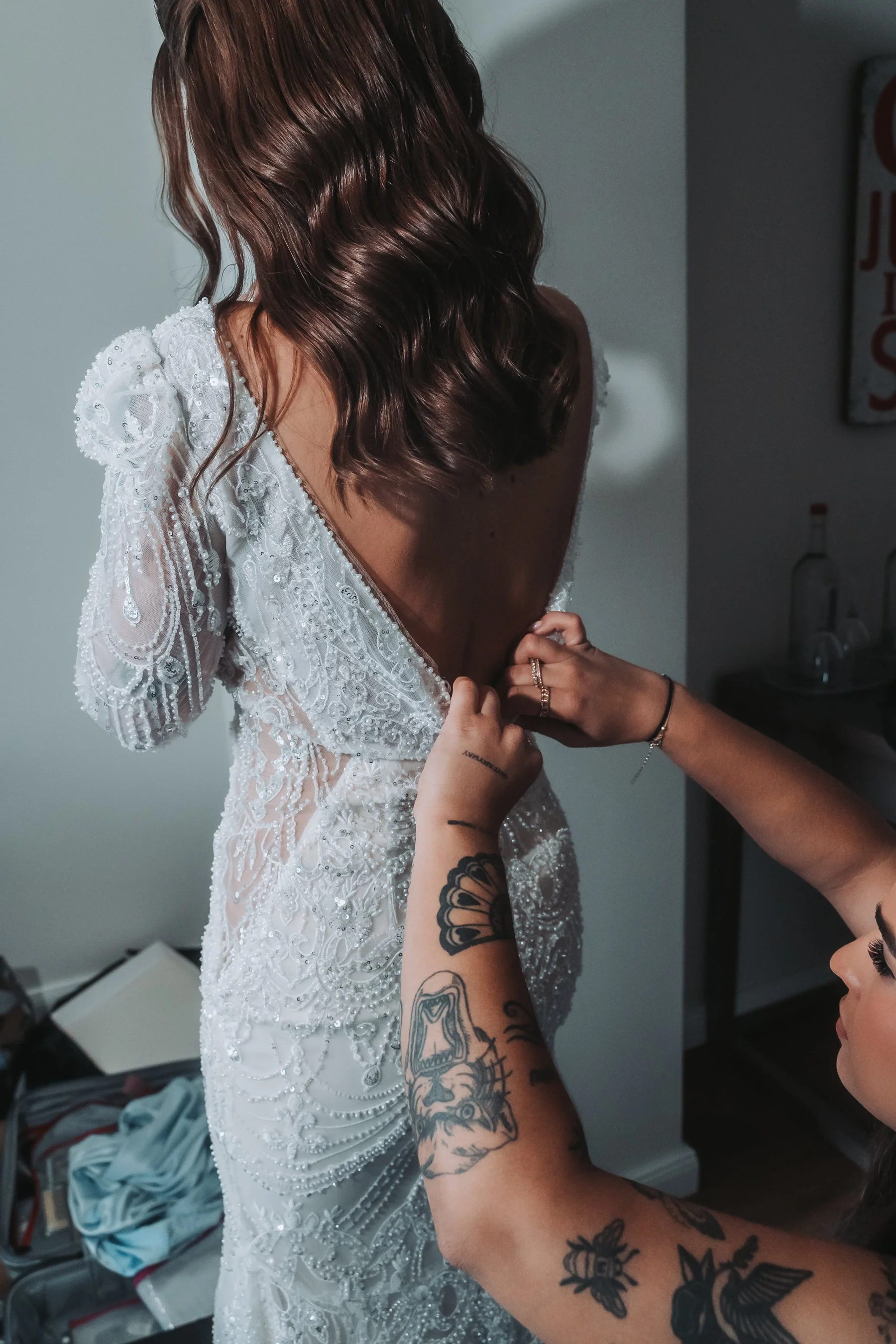 A woman in a white, intricately beaded dress with beadwork details adjusts the back of her wedding gown, which features a low back and cap sleeves. She has wavy brown hair and is surrounded by tattooed arms helping her.