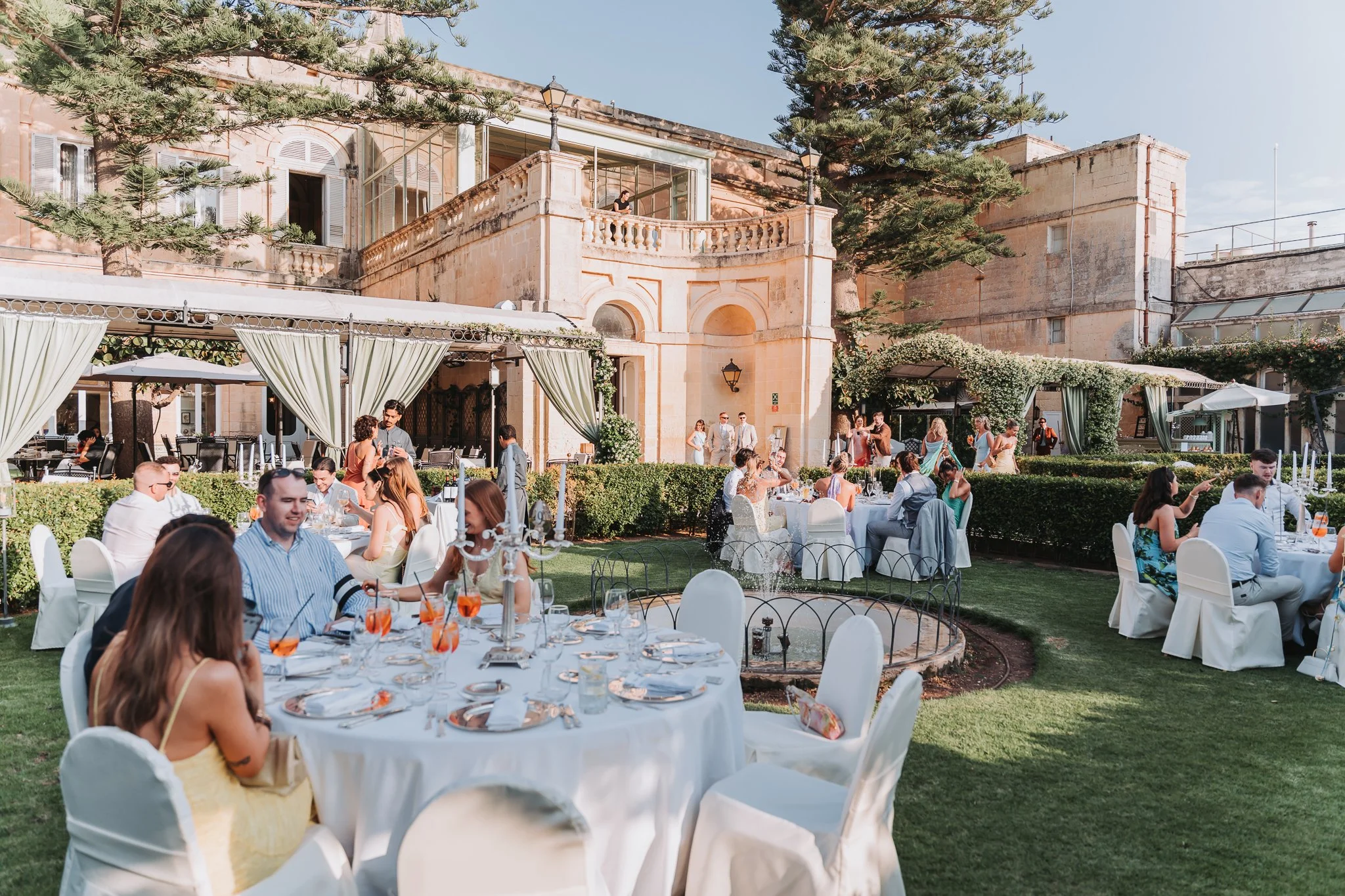 People attending an outdoor wedding reception or celebration in a courtyard with elegant white tables, chairs, and floral decorations, featuring a historic building with a balcony and large trees in the background.