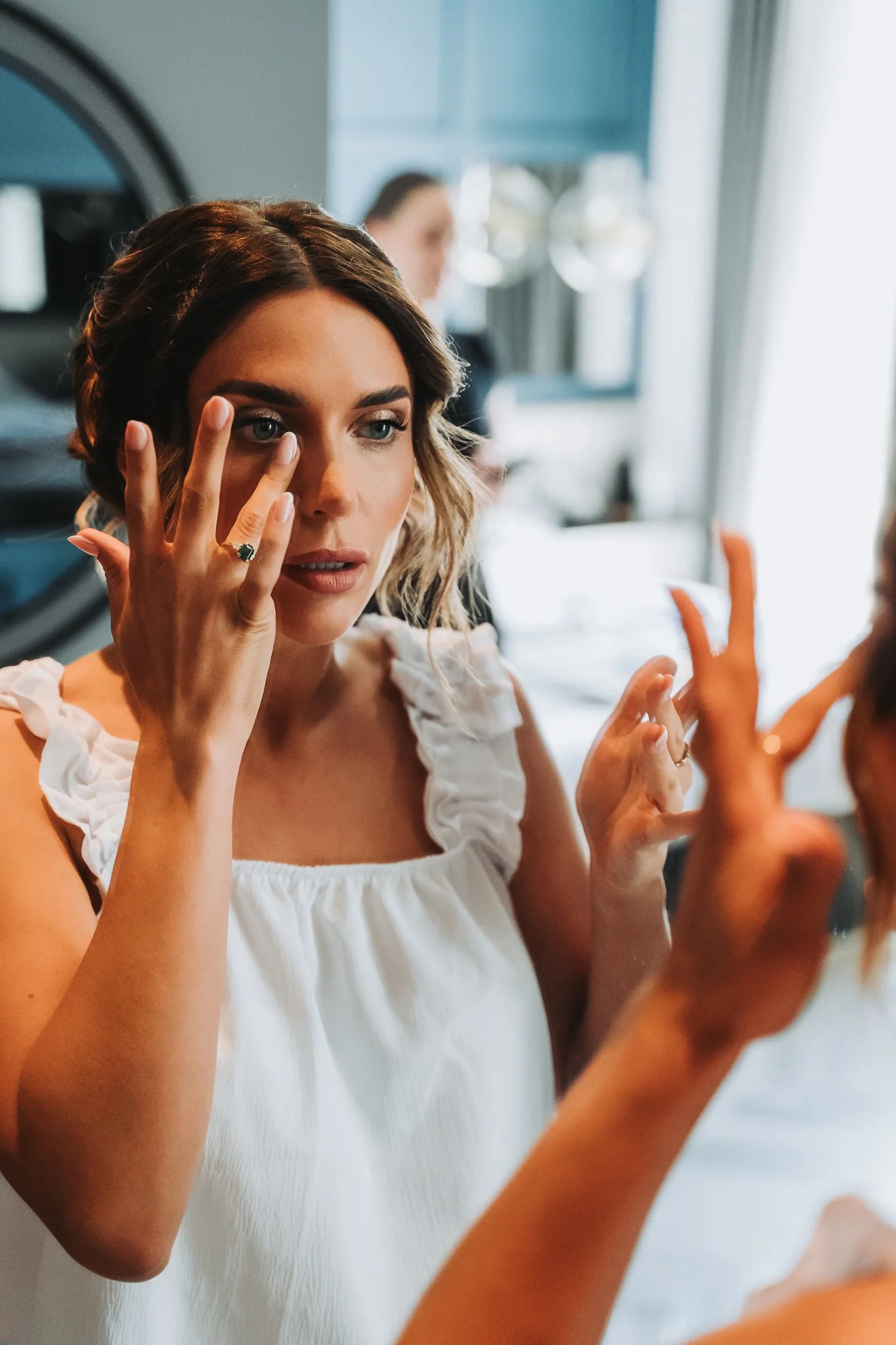 A woman with wavy hair applying makeup to her face in front of a mirror, with a person in the background in a brightly lit room.