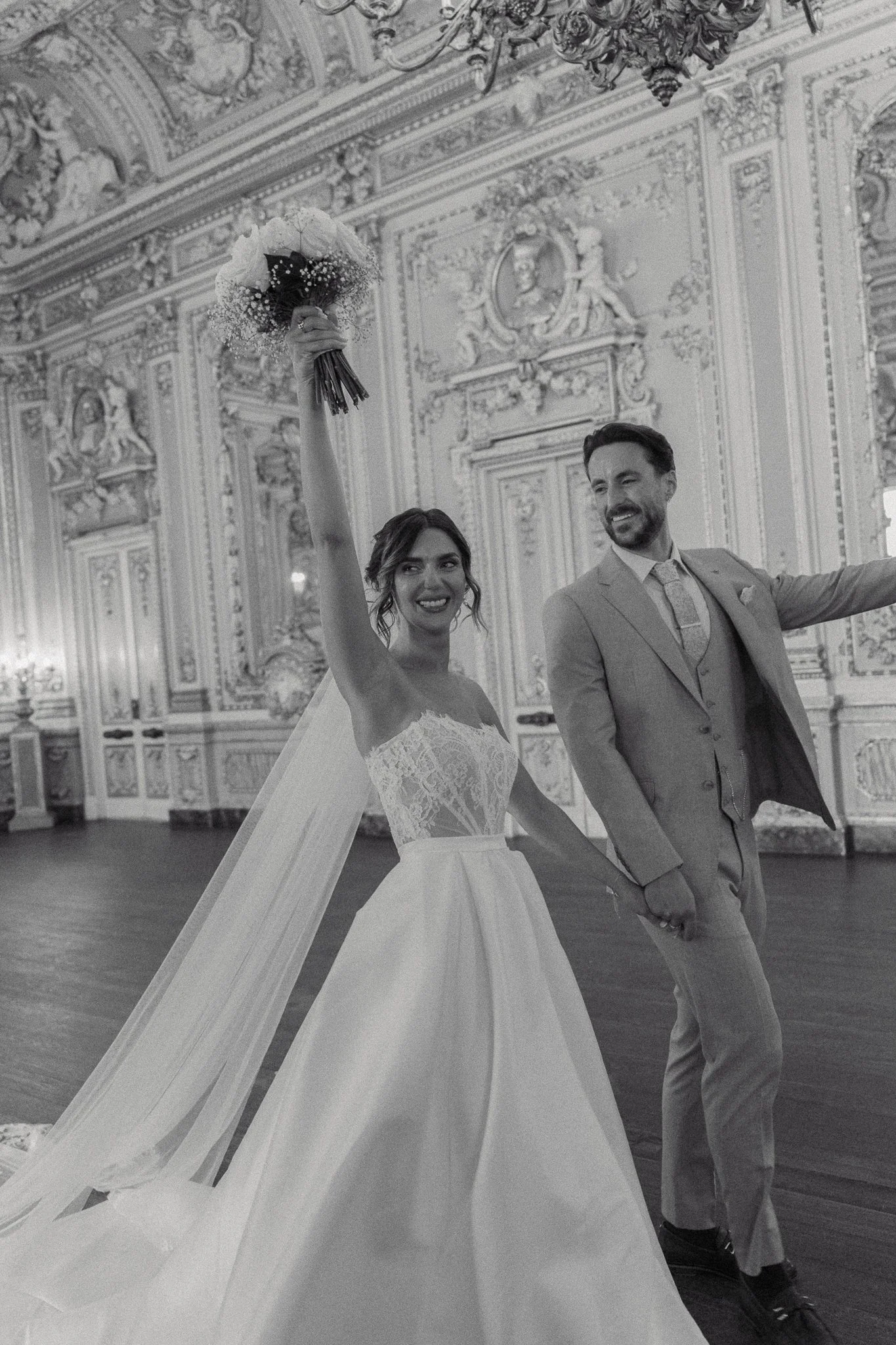A bride and groom celebrating at their wedding in an ornate hall, with the bride holding a bouquet raised in the air and the groom smiling.