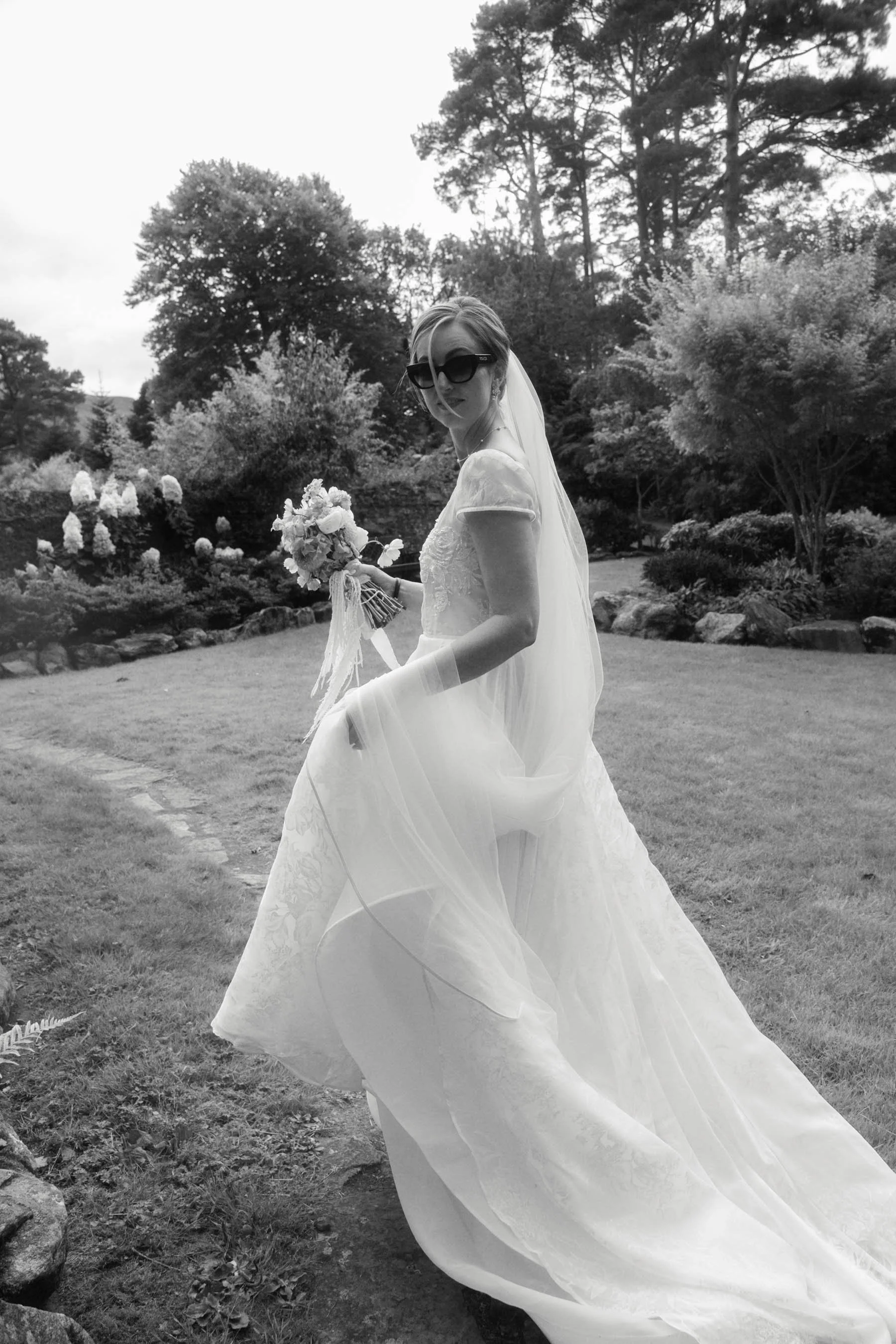 A woman in a white wedding dress holding a bouquet of flowers, wearing large sunglasses, standing outdoors in a garden with trees and shrubs, captured in black and white.