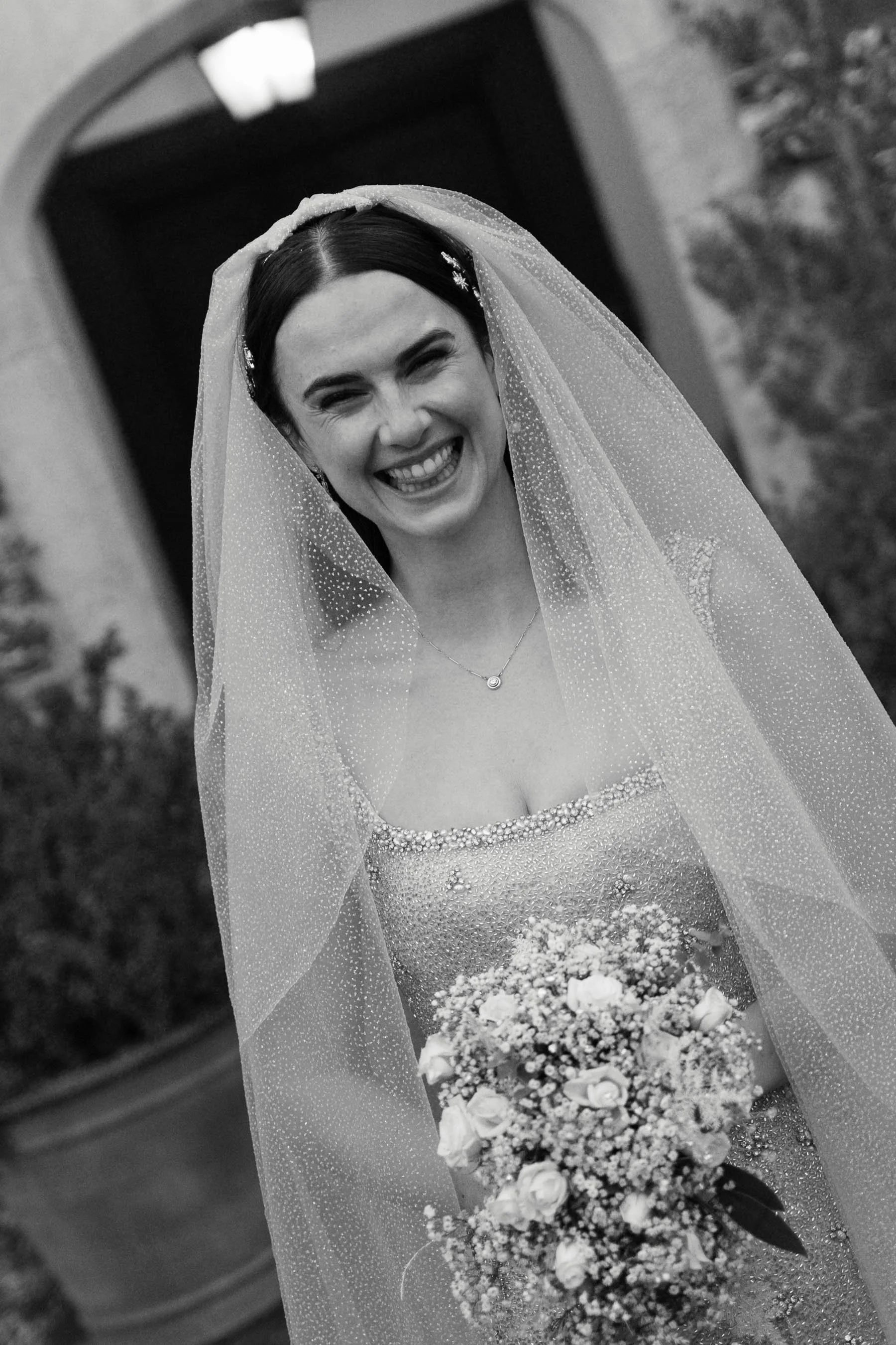 Black and white photo of a smiling bride with a veil, holding a bouquet of flowers, standing near a door with stone walls.