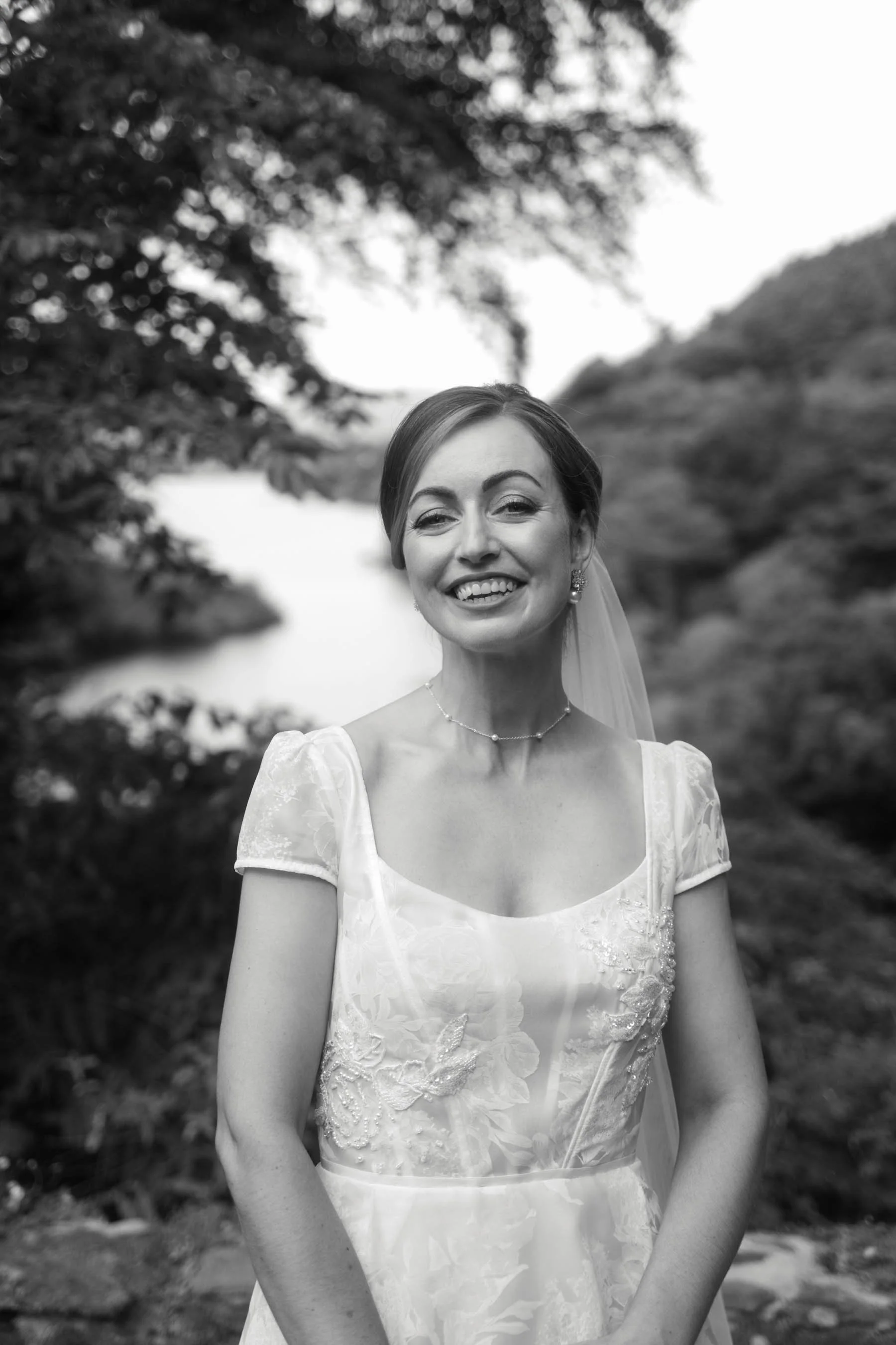 A smiling woman in a wedding dress outdoors with trees and hills in the background.