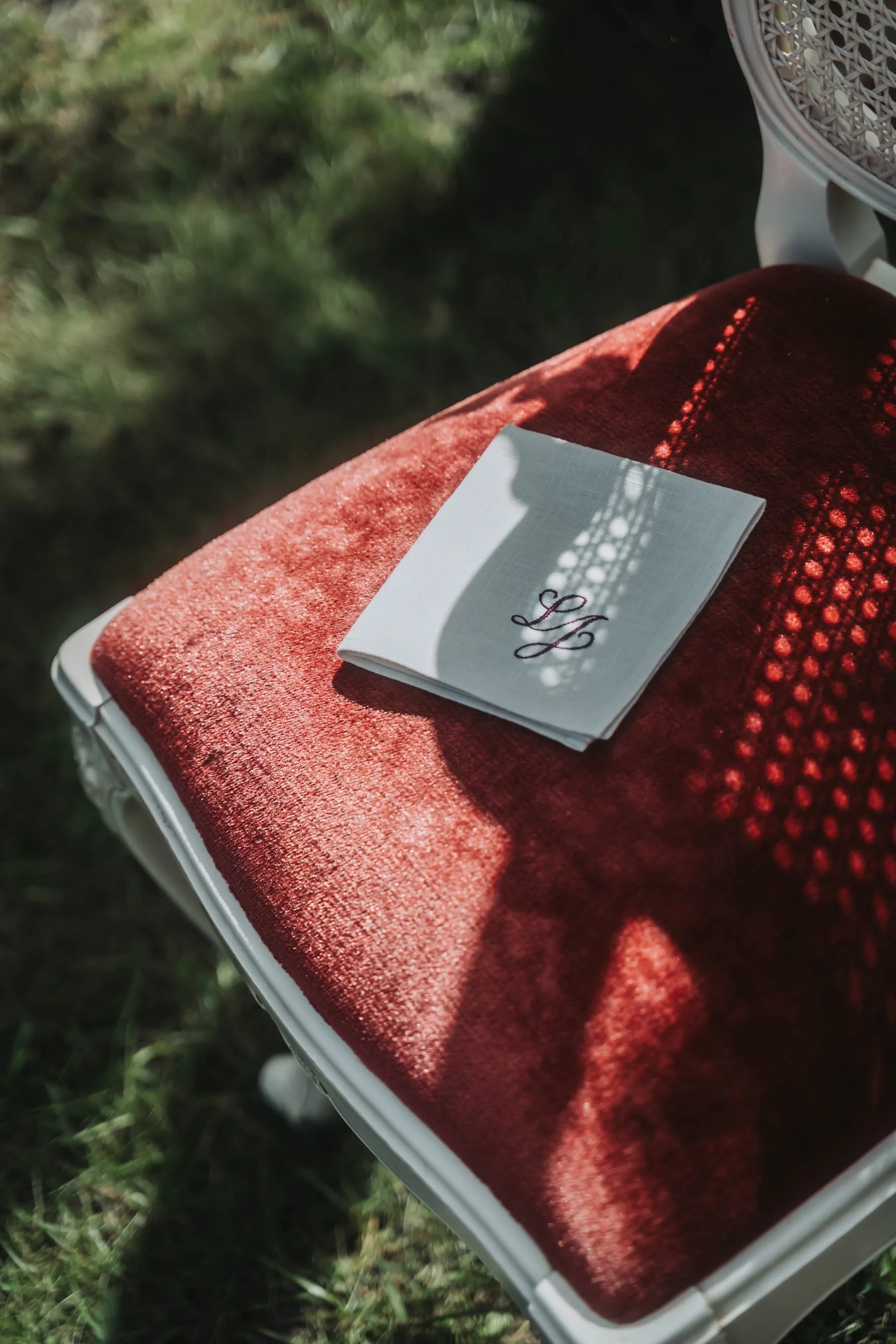 A small white bench with a red velvet cushion on top, a white cloth with embroidered initials 'LY,' and a white fan, placed on green grass, with shadows casting on the cloth.