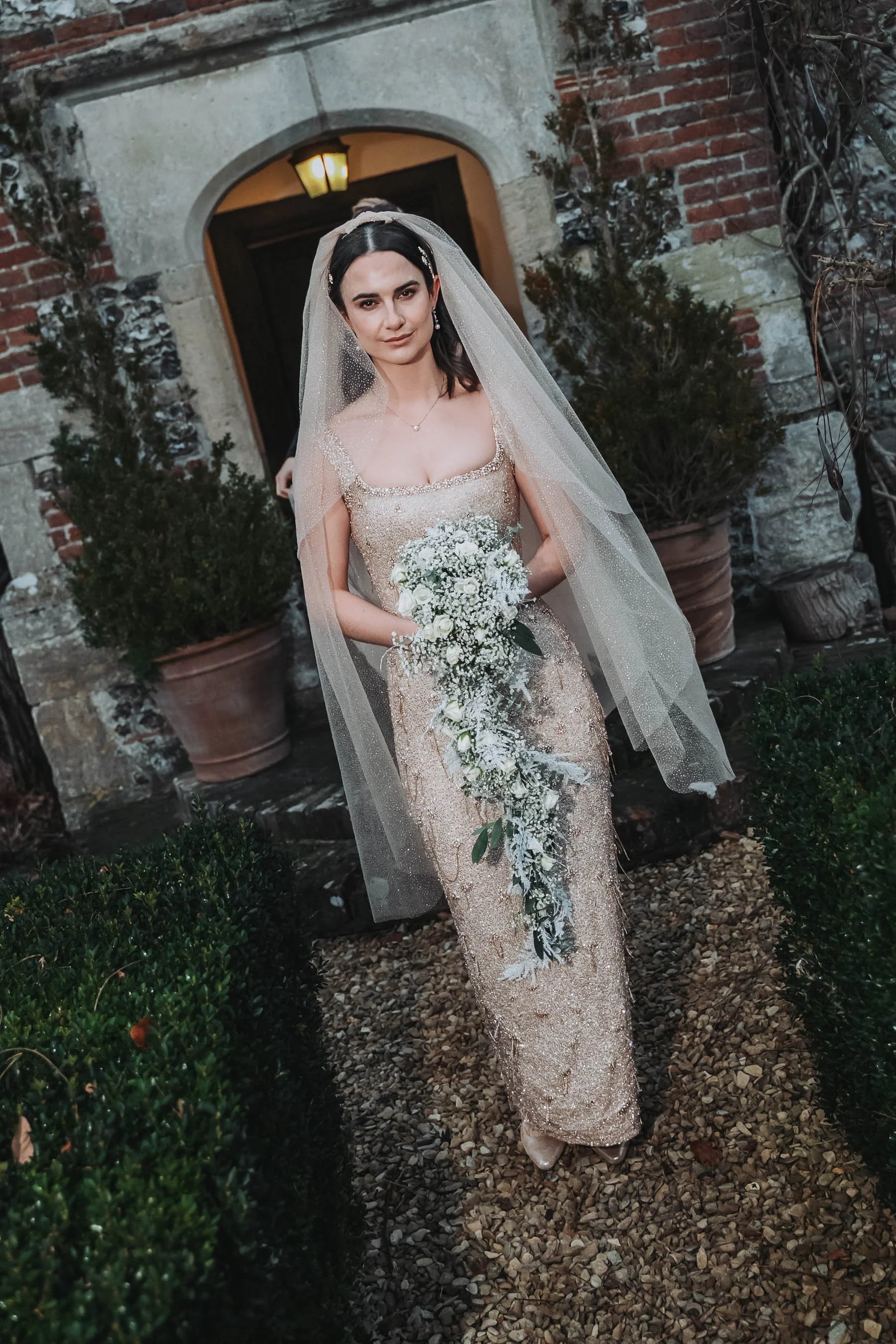A bride in a sparkling gold gown with a sheer veil standing outdoors in front of a brick building with potted greenery, holding a cascading bouquet of white flowers and greenery.