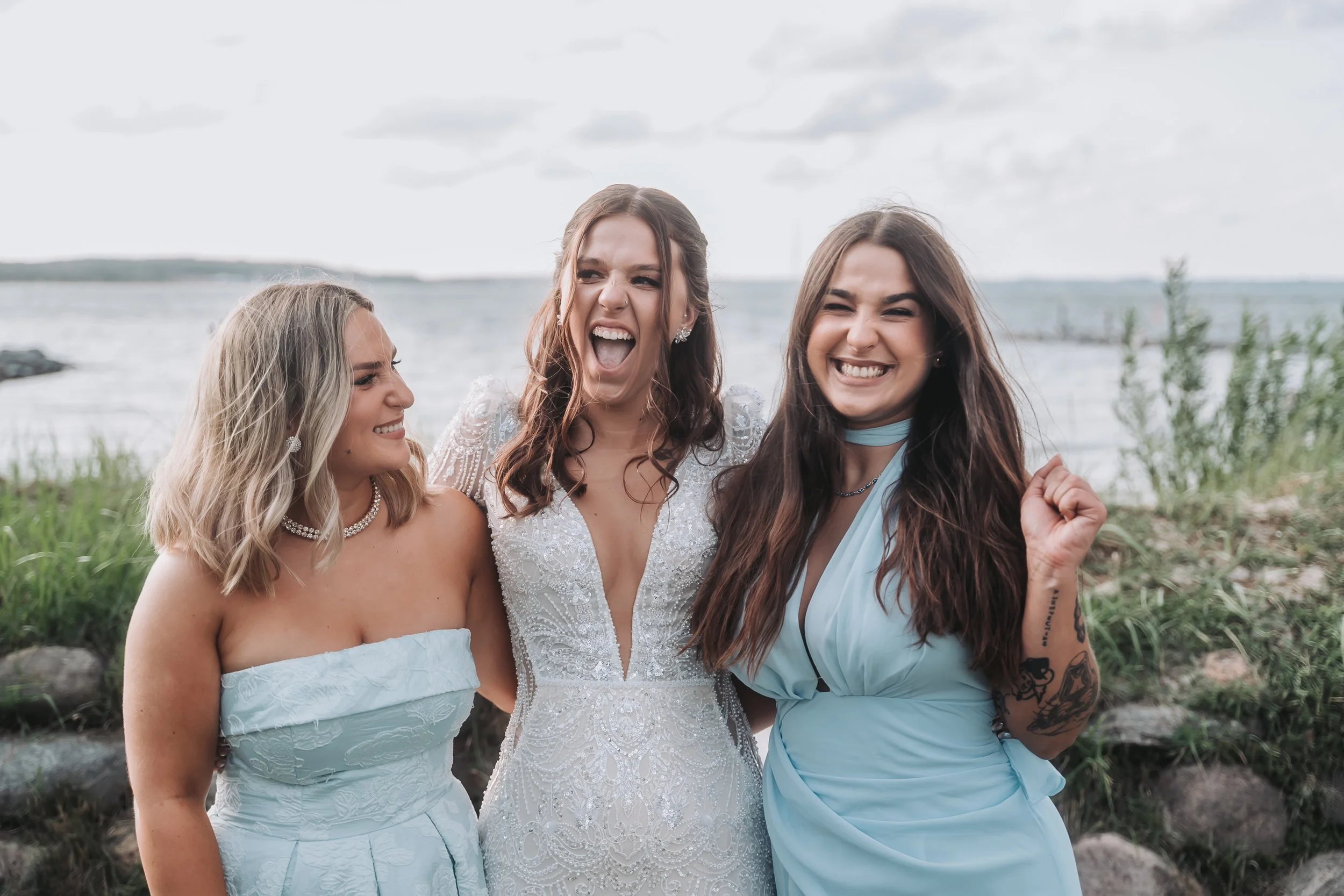 Three smiling young women standing together outdoors near a body of water with grassy area and cloudy sky in the background, celebrating and enjoying each other's company.