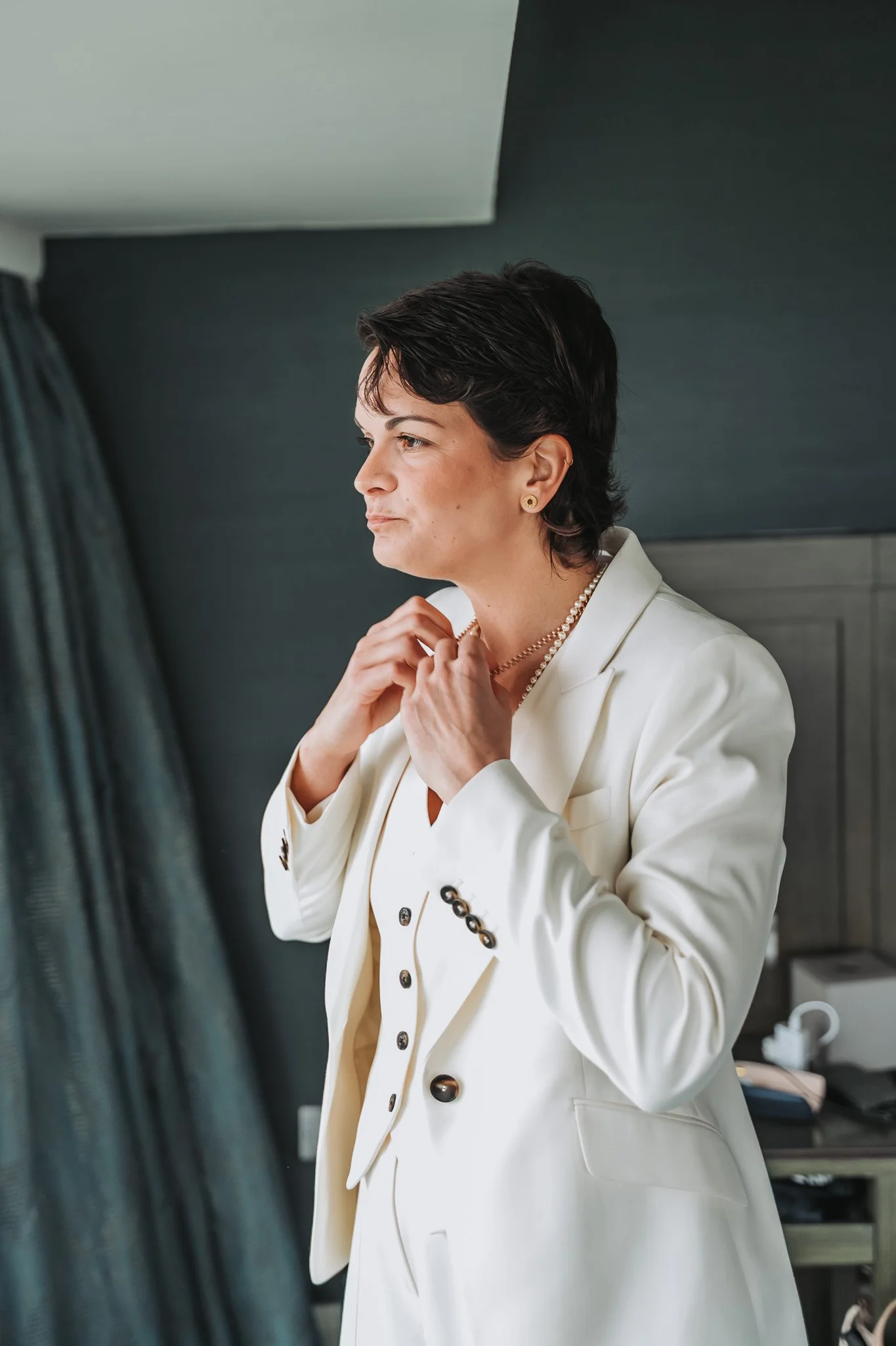 A woman with short dark hair wearing a white blazer and pearl jewelry, adjusting her collar in a room with dark walls and curtains.