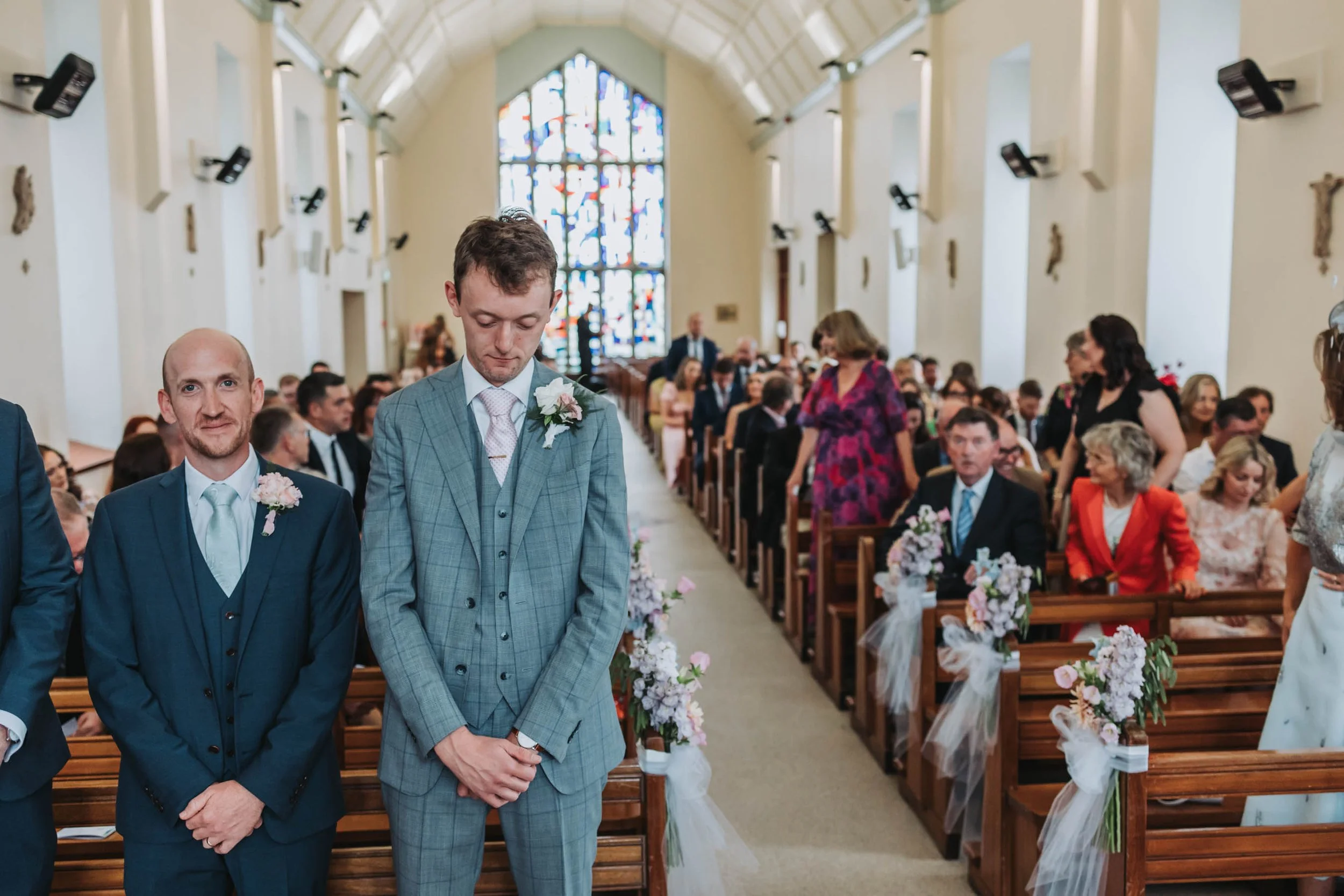Two men standing in a church during a wedding ceremony, with guests seated in the background and stained glass windows behind them.