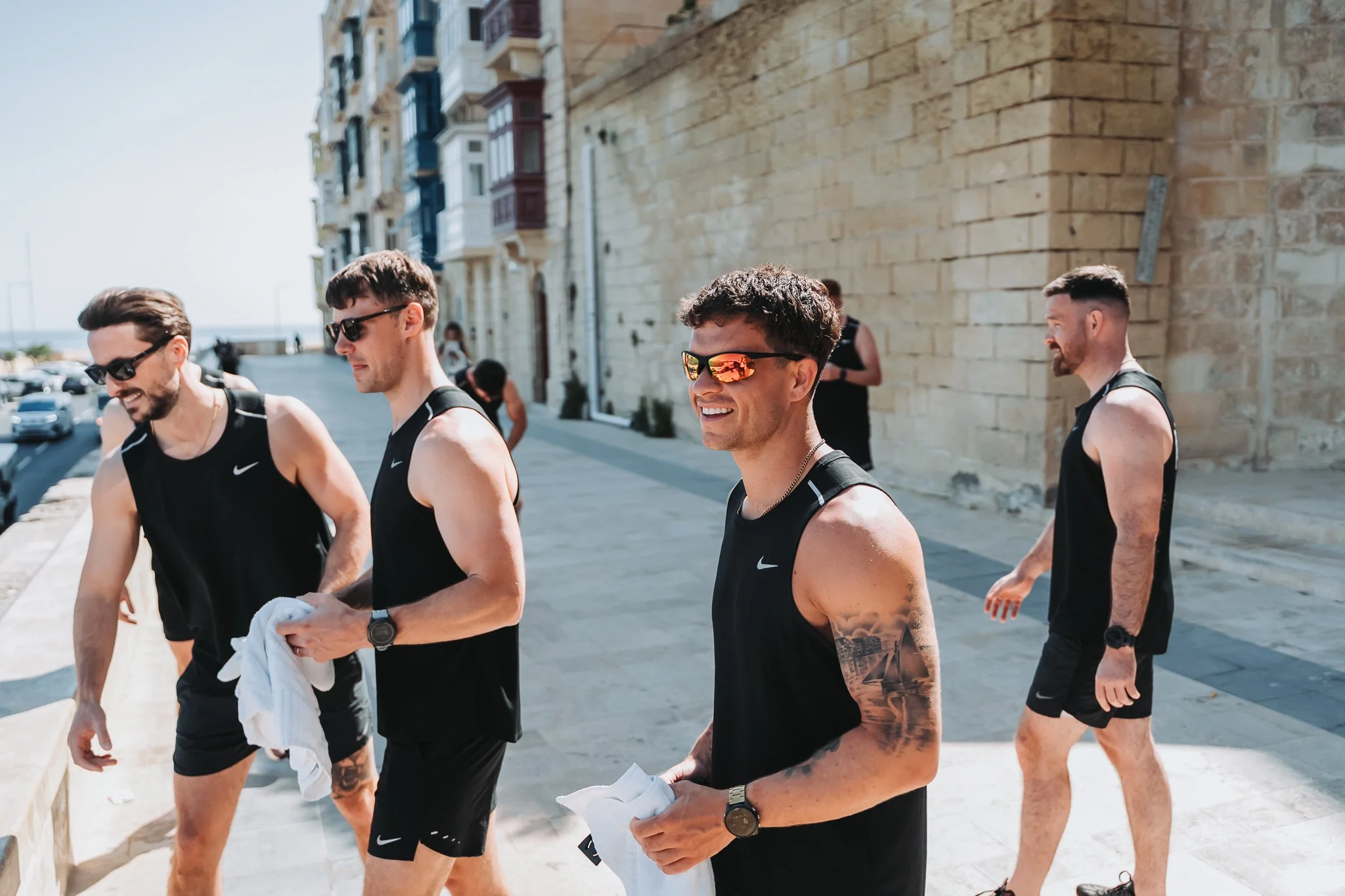 Four men in black athletic clothing walking outdoors on a sunny day with a stone building in the background.
