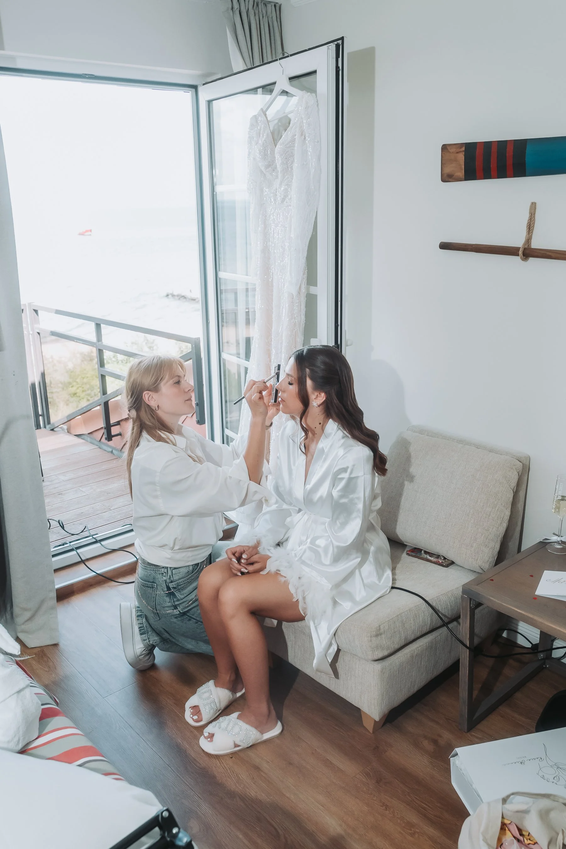 Two women preparing a bride for her wedding, one applying makeup and the other seated in a satin robe, in a room with a large window and ocean view.