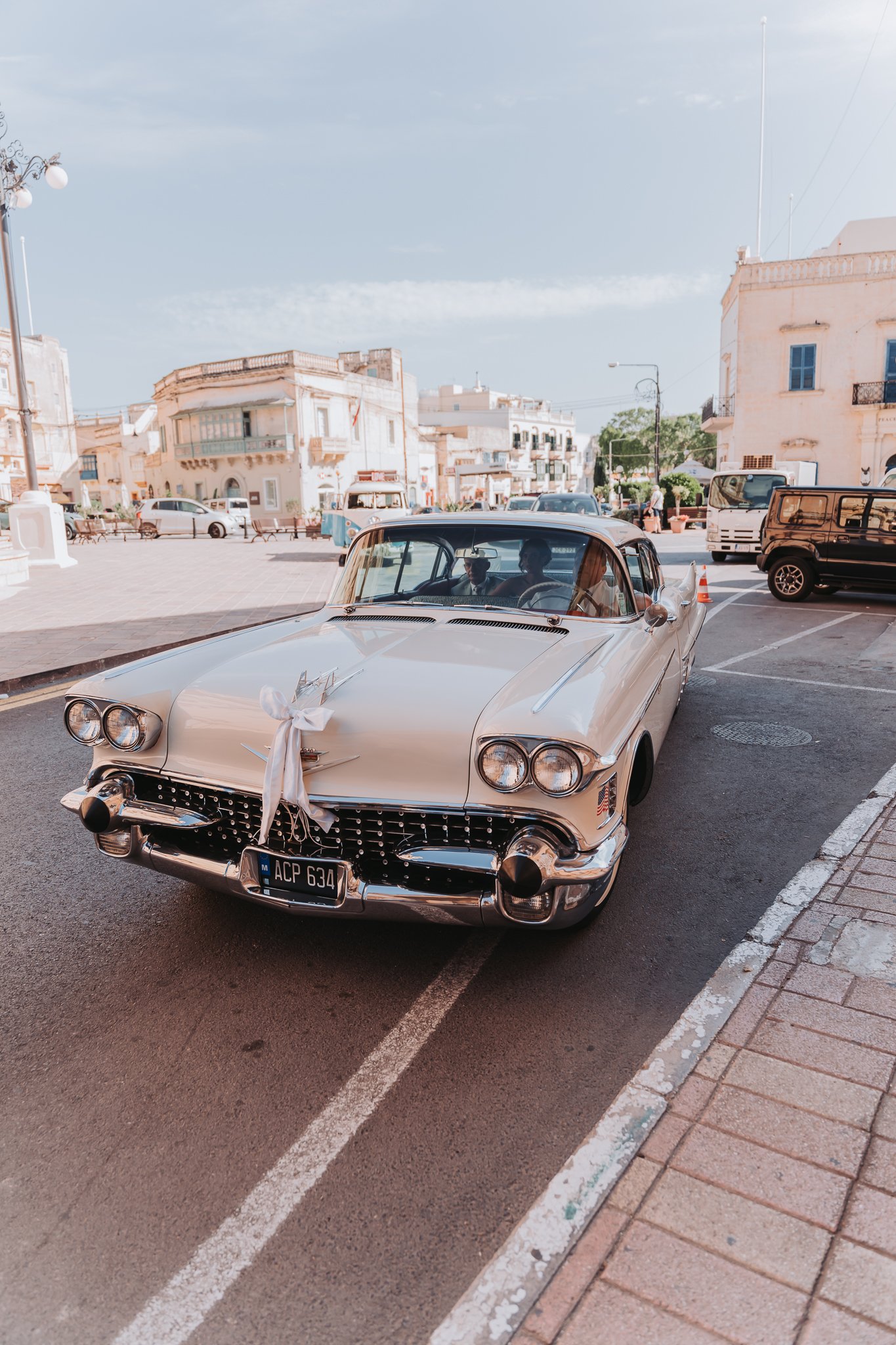 A vintage white Cadillac car parked on a street with other vehicles and old buildings in the background, under a partly cloudy sky.