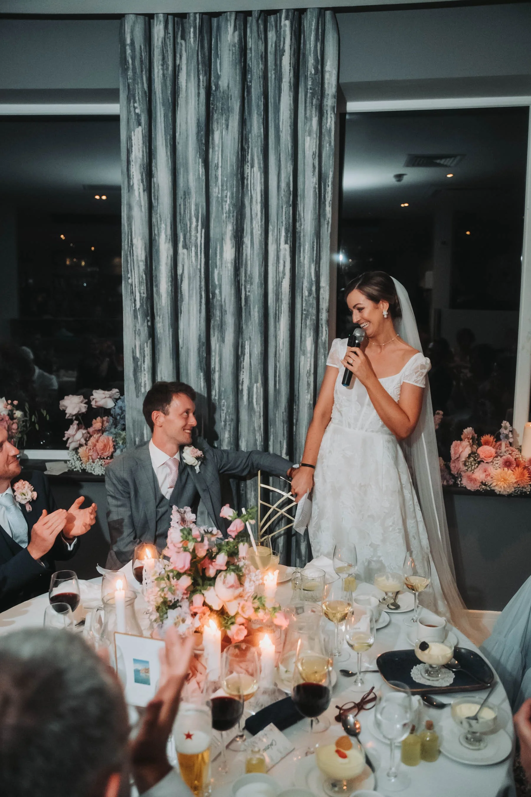 A bride giving a speech holding a microphone at a wedding reception, seated at a table decorated with flowers and candles, surrounded by guests in formal attire.