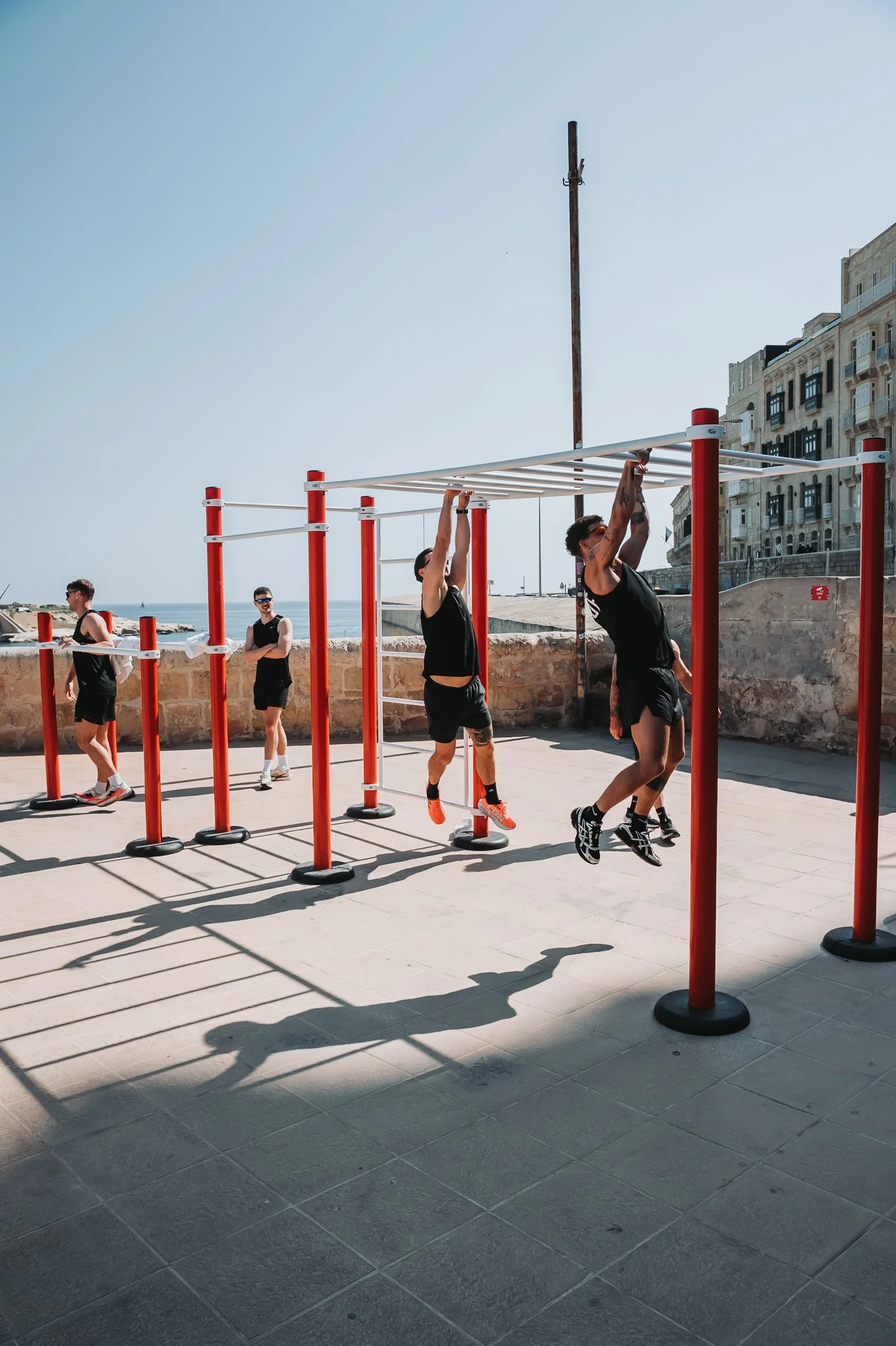 Four men in black athletic clothing and sunglasses exercising outside near the beach, using outdoor workout equipment with red and white bars, against a backdrop of historic buildings and a clear blue sky.