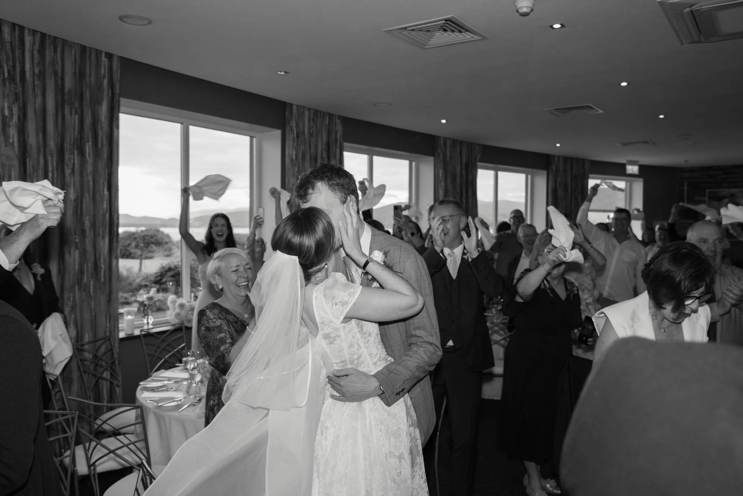 A black-and-white photo of a wedding reception with a bride and groom kissing. Guests around them celebrate and throw napkins in the air. The event is indoors with large windows and scenic outdoor views.