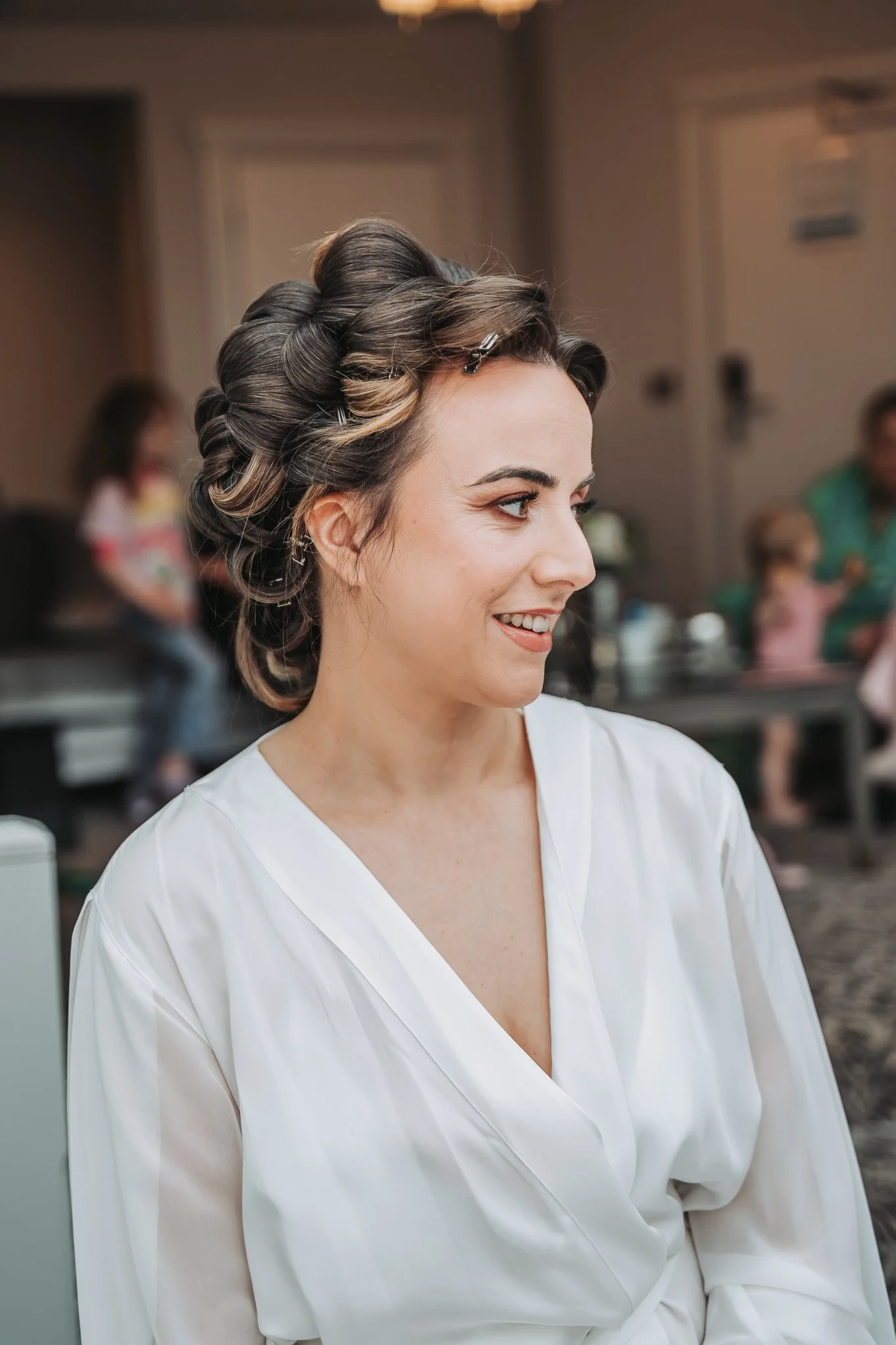 A woman with dark hair styled in vintage rollers wearing a white robe, smiling and looking to the side in a salon or dressing room setting with blurred background of other people and furnishings.
