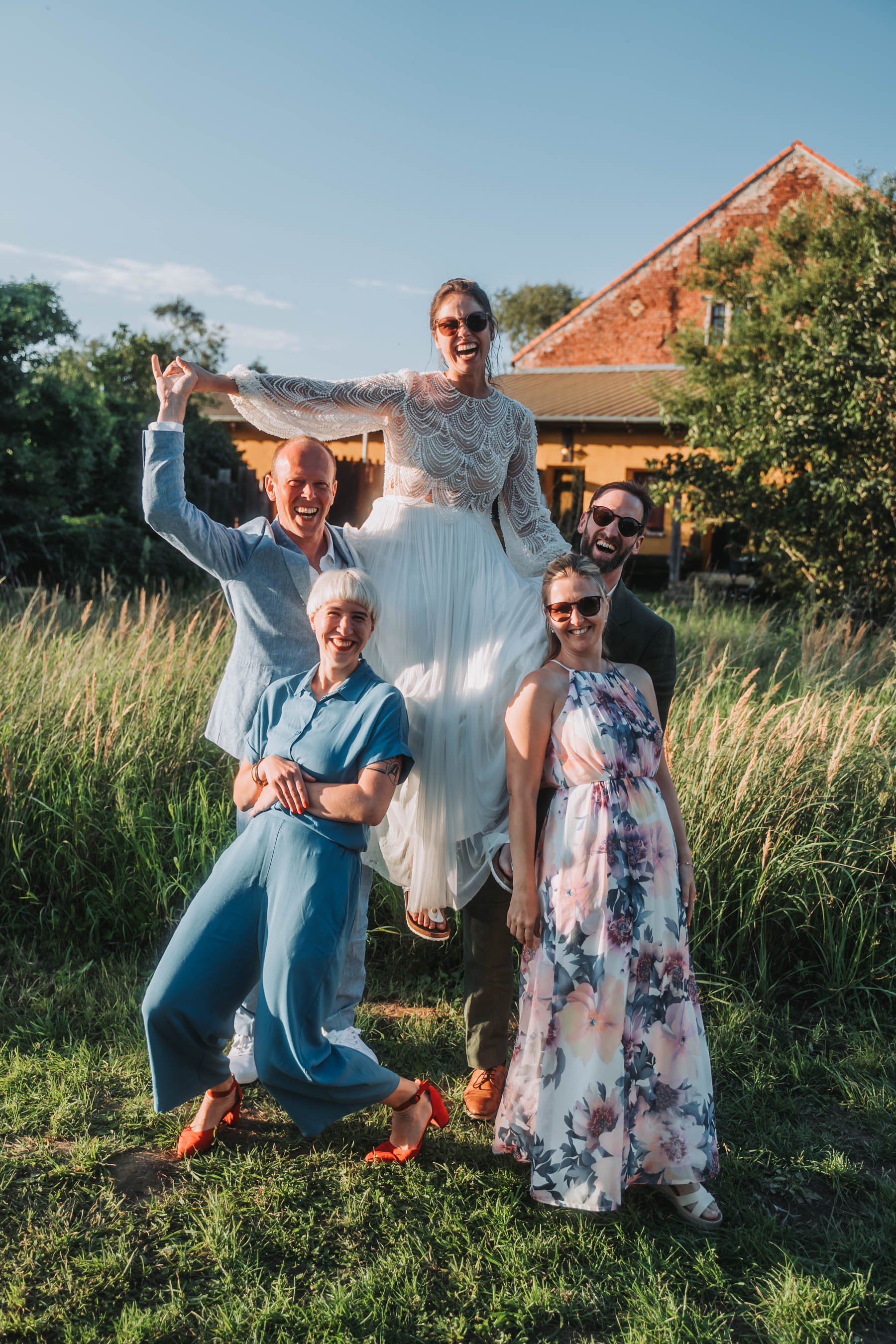 A group of five people outdoor in a grassy field, with a woman in a white dress being lifted by four others, all smiling and laughing, under a clear blue sky with a red brick house and trees in the background.