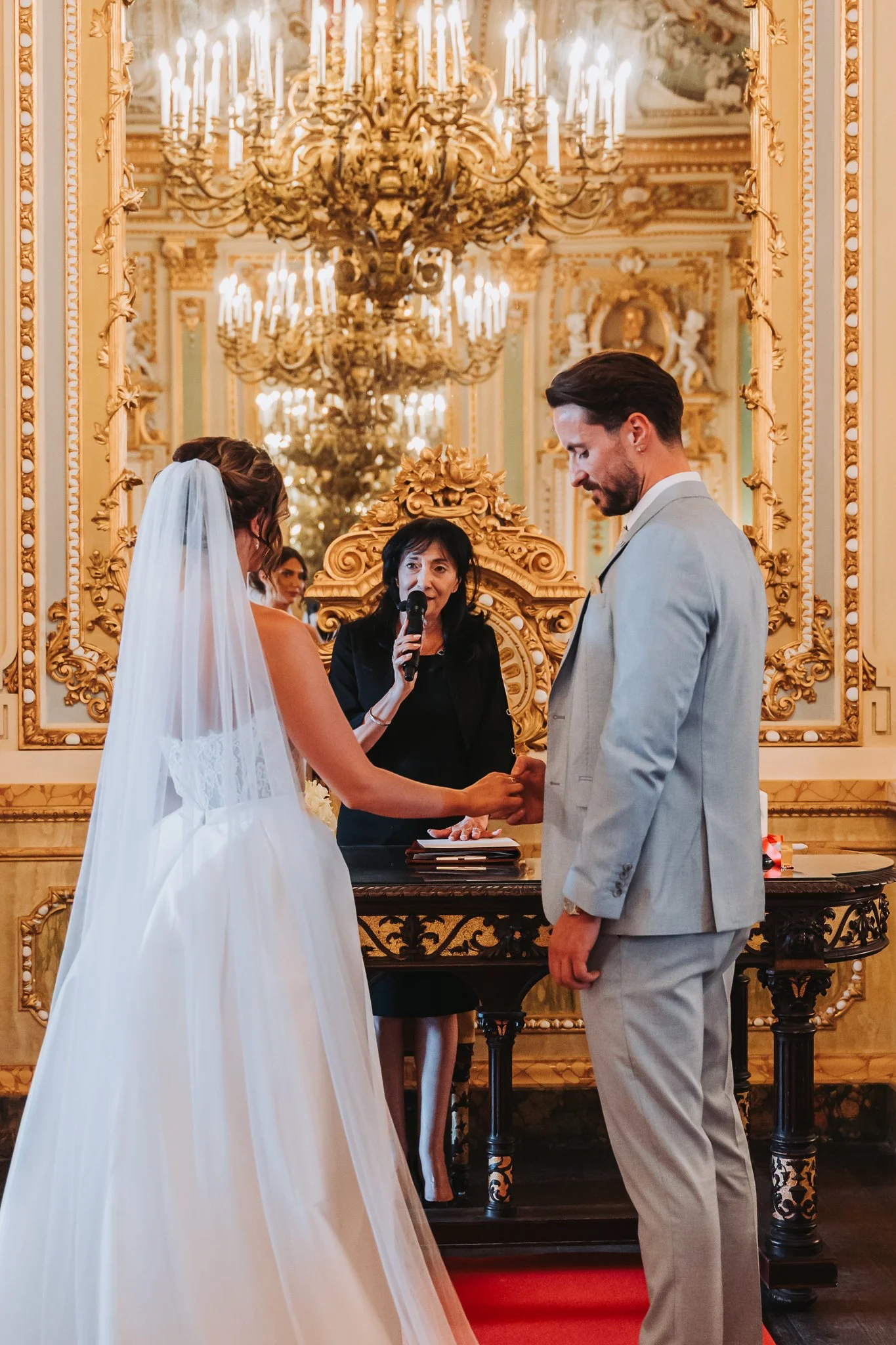 A wedding ceremony with a bride and groom holding hands facing each other, with an officiant speaking into a microphone, in an ornate, gold-decorated room featuring grand chandeliers.