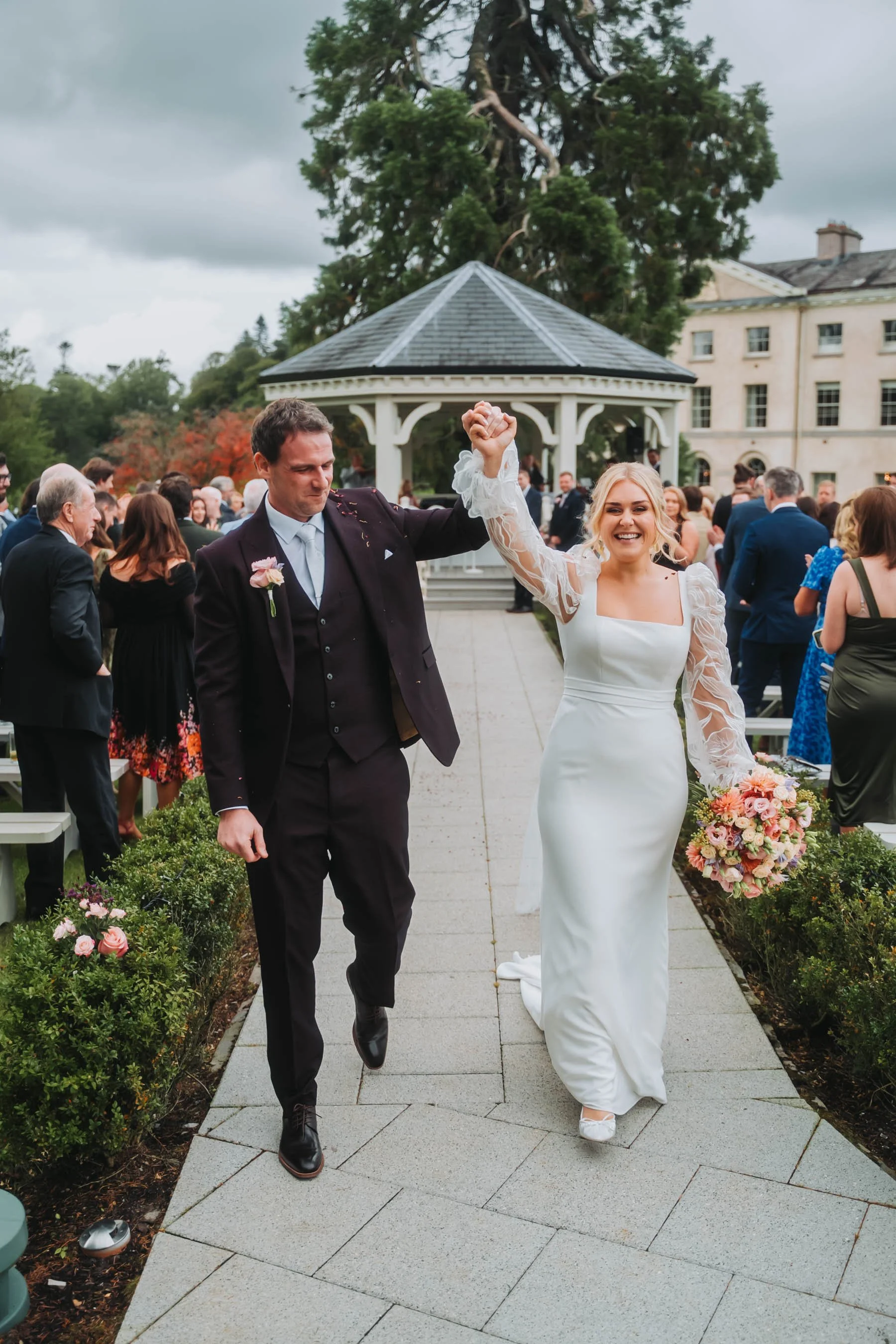 A newlywed couple is walking outdoors on a paved pathway, holding hands and smiling. The bride is in a white wedding dress holding a bouquet of flowers, and the groom is in a dark suit. They are surrounded by guests, trees, and a building with a gaze