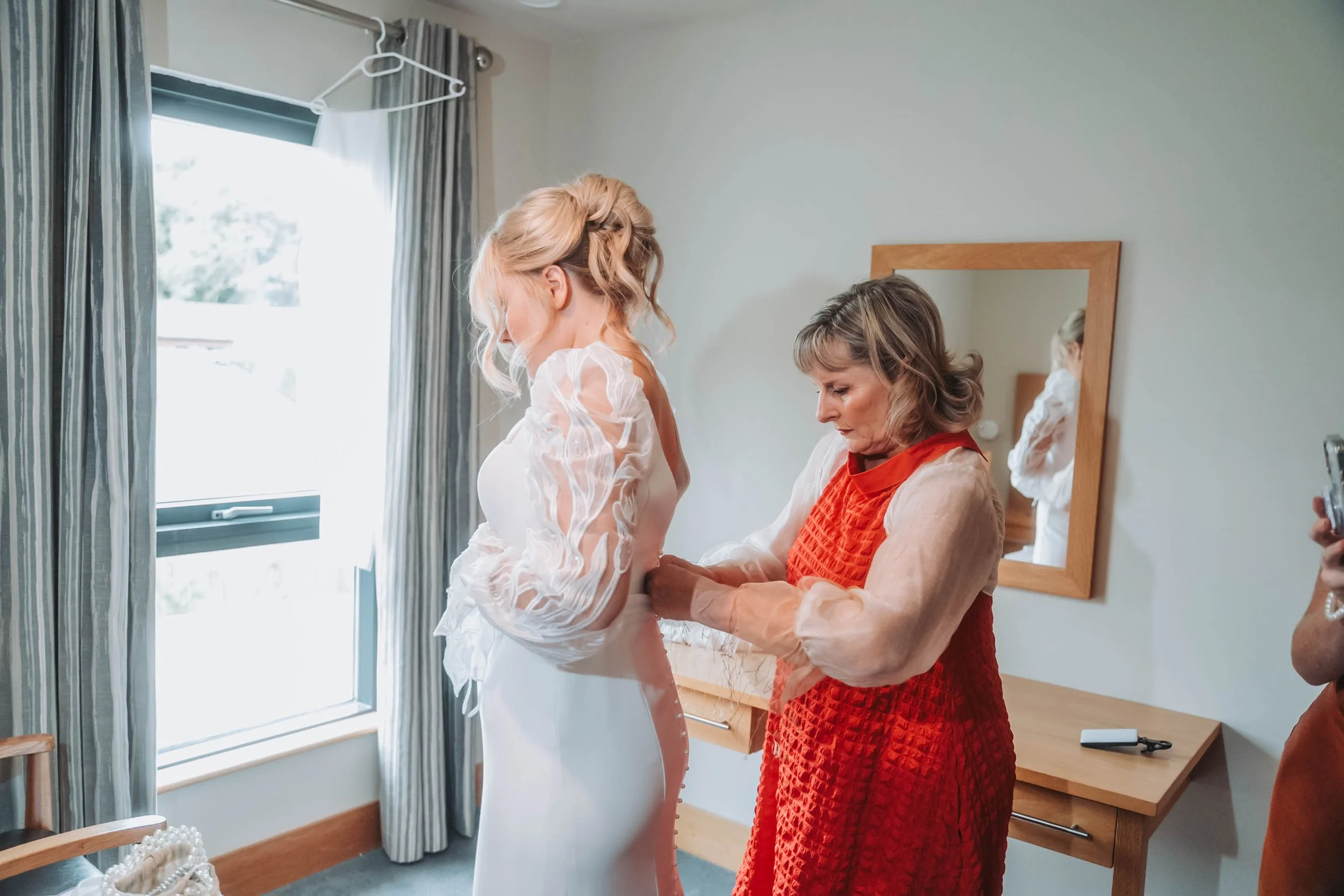 Woman in a red dress helping a bride put on a wedding dress near a window with curtains, with a mirror in the background.