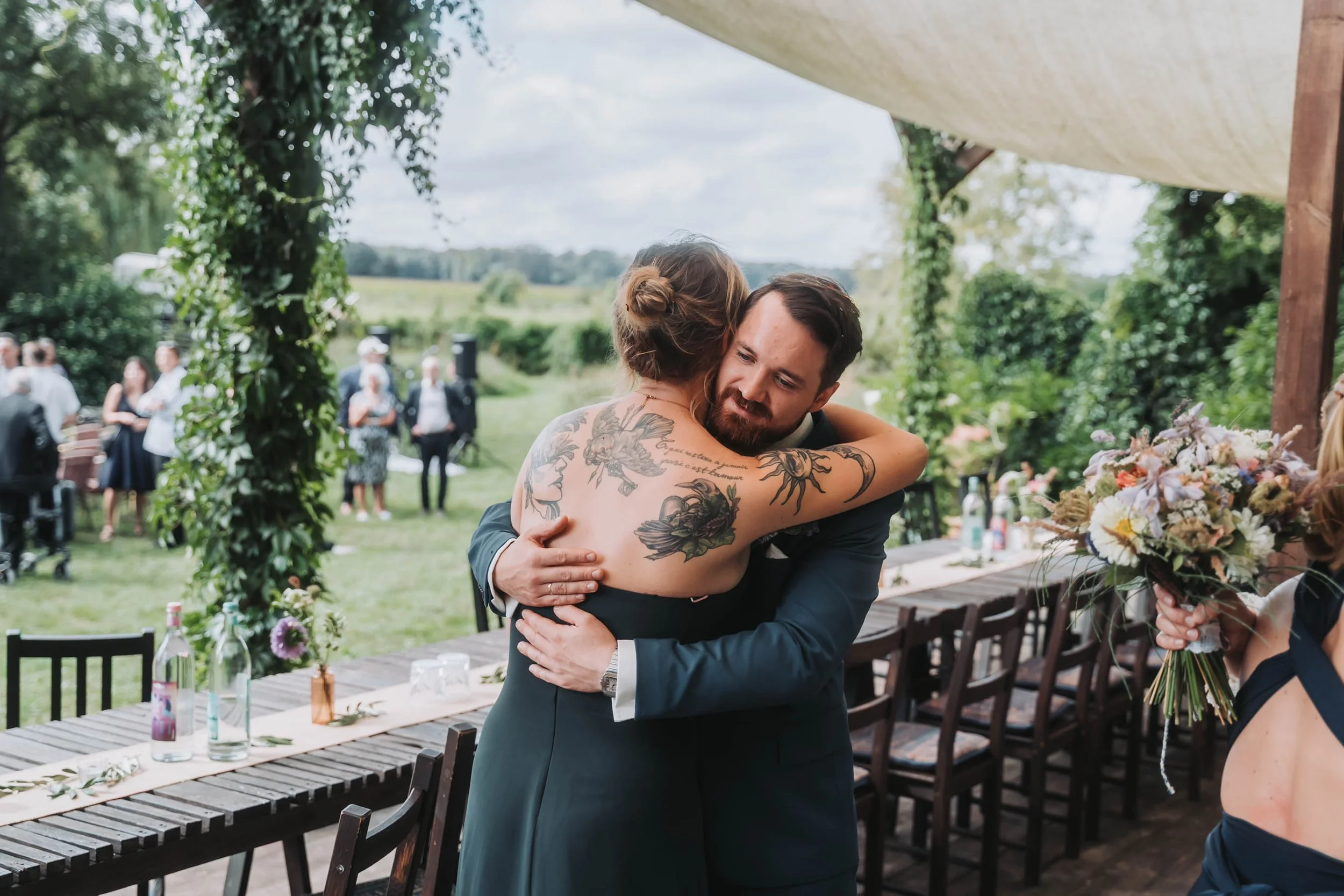 A man and woman embrace at an outdoor wedding reception, with guests in the background and a scenic green landscape.