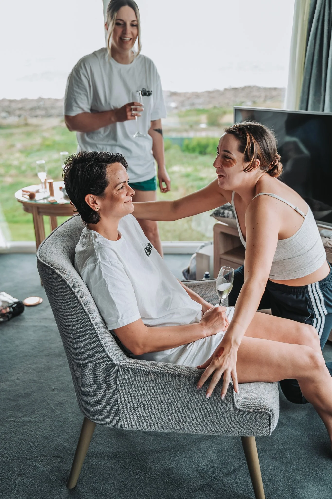 A woman with tattoos and a woman with short dark hair and a black eye are sitting together on a gray armchair, smiling and holding a glass of champagne. A woman with long blonde hair, wearing a white t-shirt, is standing behind them, smiling and hold